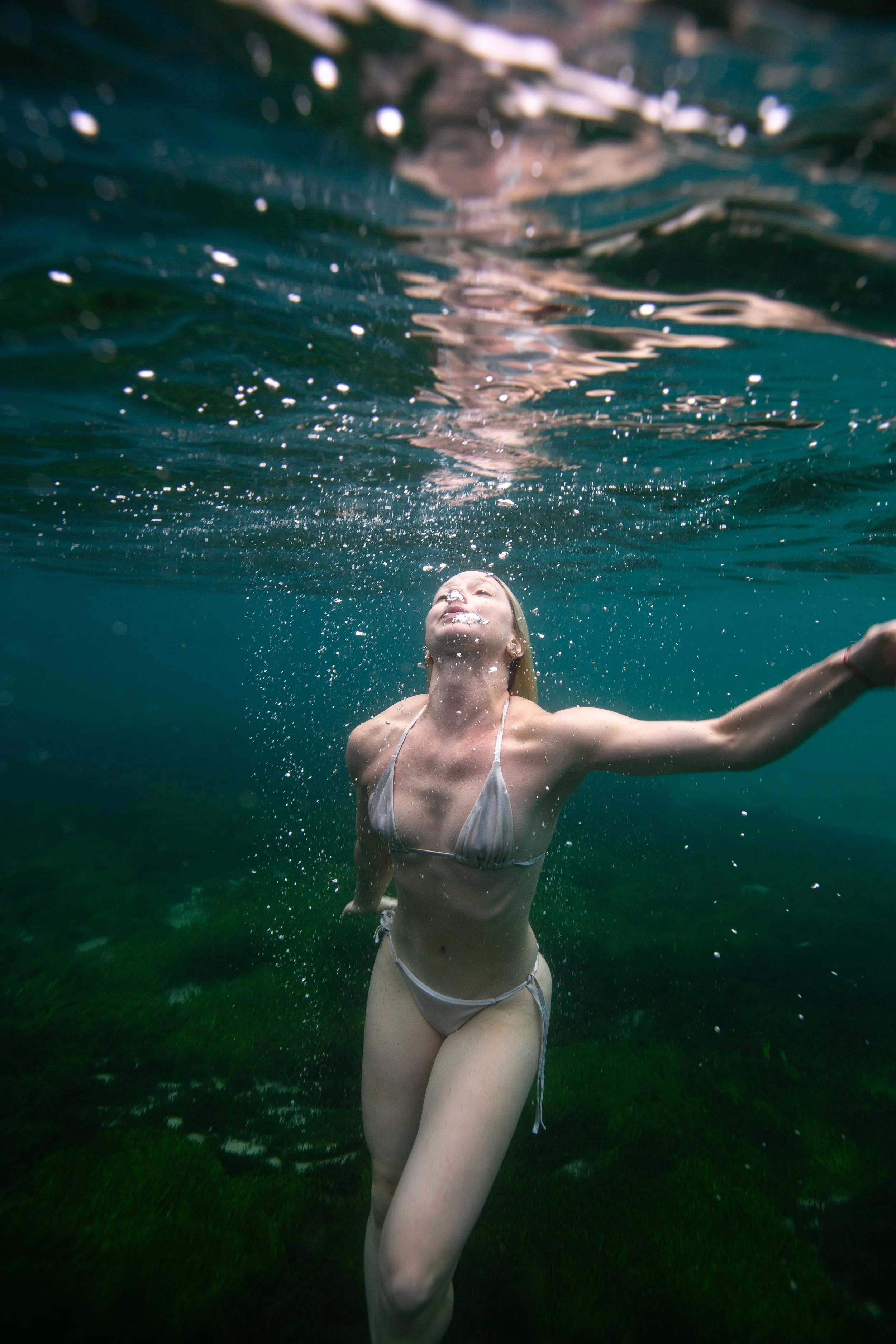A woman in a bikini underwater, arms outstretched, with the water surface above her and a greenish underwater environment below.