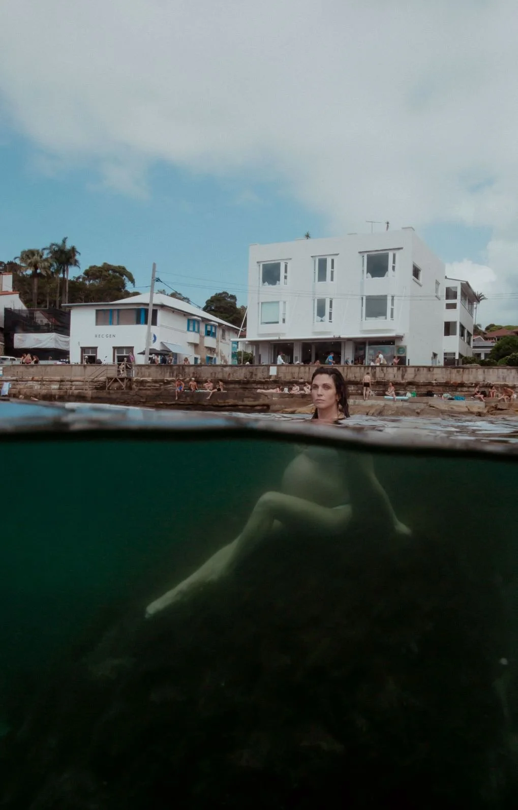 A woman swimming in a body of water near a shoreline with houses and buildings in the background, and people on the rocks and shoreline.