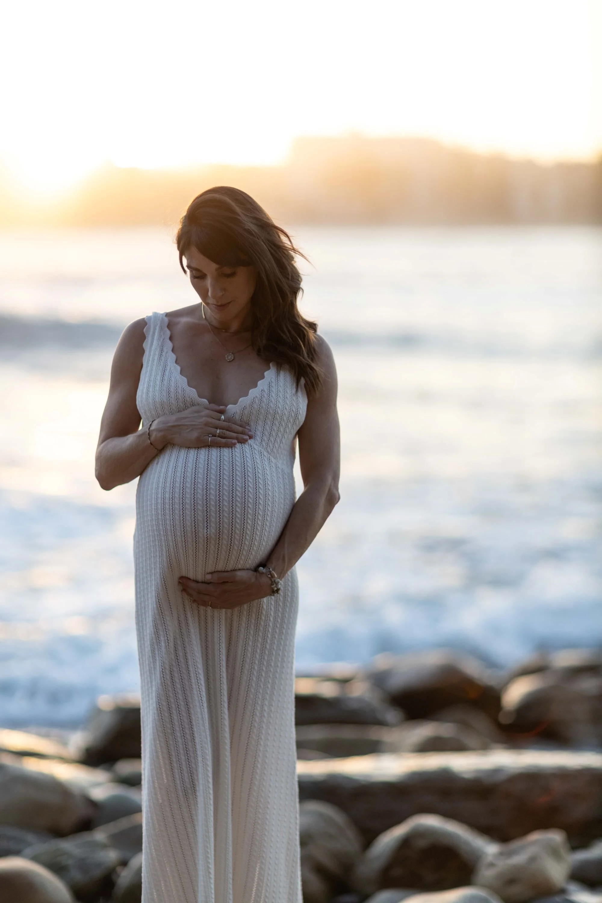 A pregnant woman in a long white dress standing on a rocky beach at sunset, with a calm ocean in the background.  Maternity style in Sydney.
