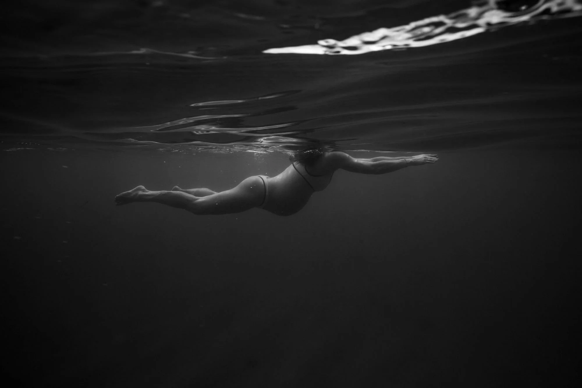 A pregnant person swimming underwater in a black and white photo with their body stretched out horizontally and arms extended forward.  Maternity style in Sydney.