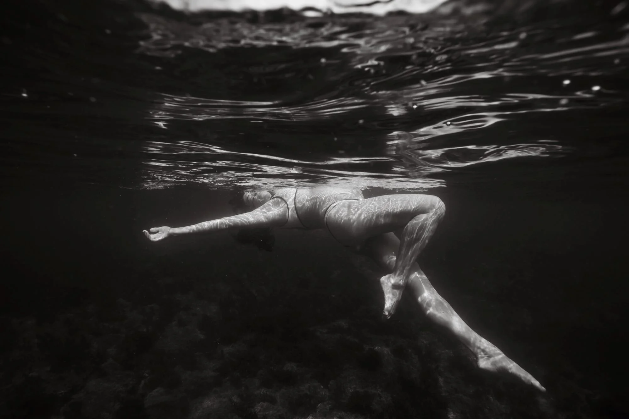 A pregnant woman person swimming underwater in a body of water, with water ripples and light reflections visible on her body.  Maternity style in Manly, Sydney.
