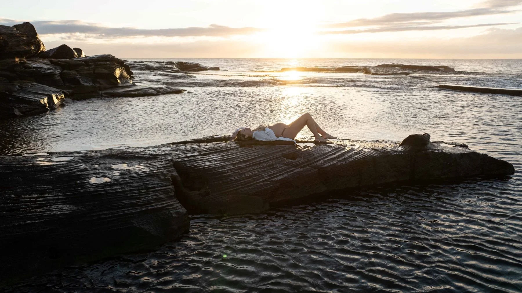 A woman lying on a large rock by the ocean at sunset, wearing a white shirt and minimal clothing, with the sun setting on the horizon and calm water surrounding her.