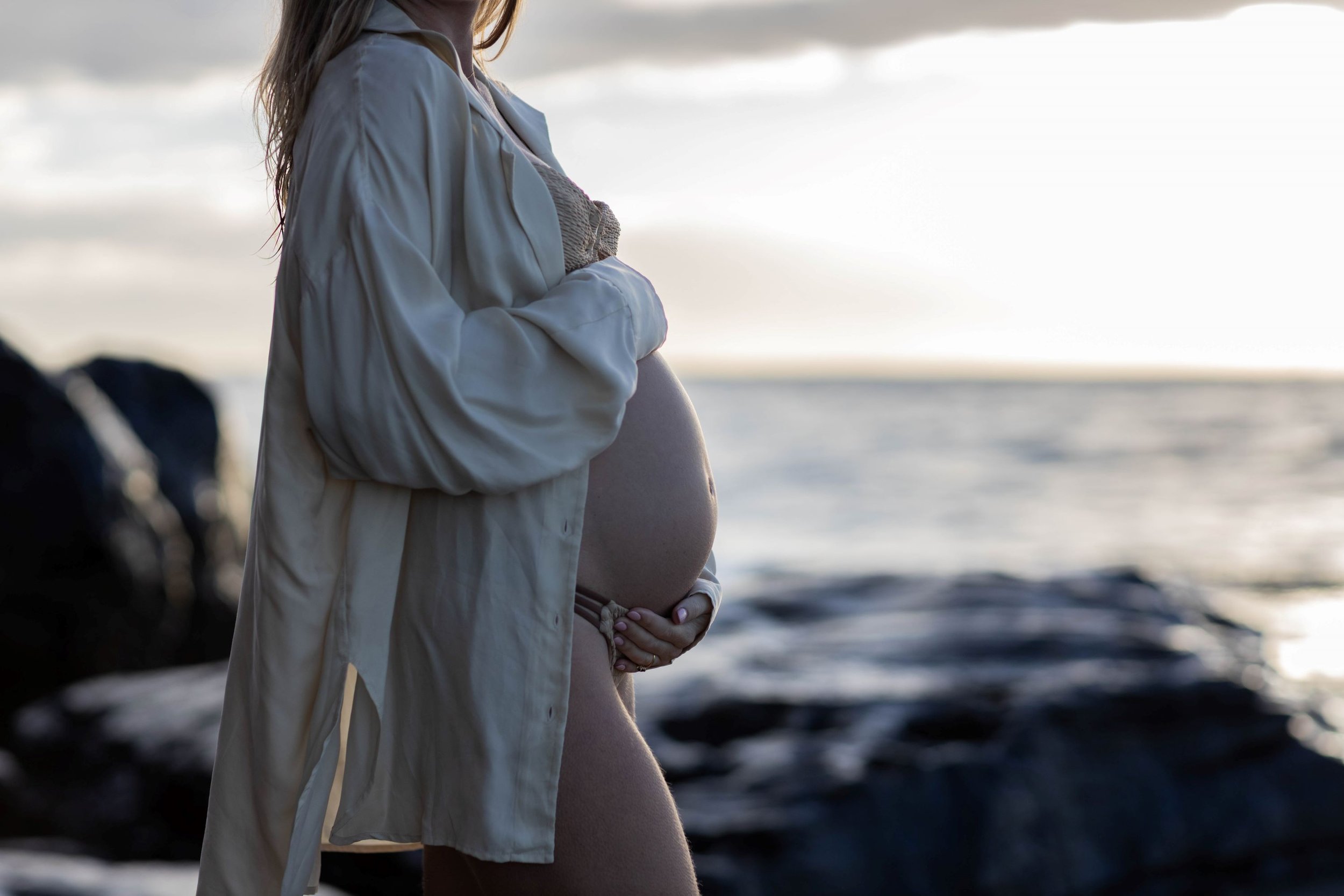 Pregnant woman standing on a beach during sunset, holding her belly with both hands, wearing a light jacket and beanie.