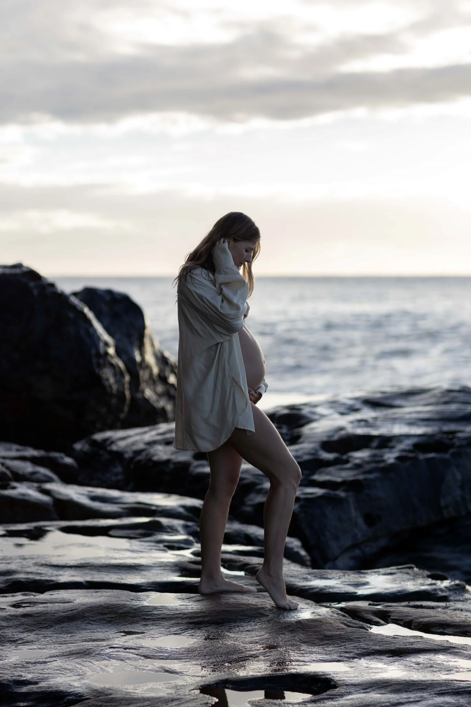 A pregnant woman standing barefoot on a rocky beach at sunset, wearing a loose white shirt, with her head bowed and hands resting on her belly, looking contemplative.