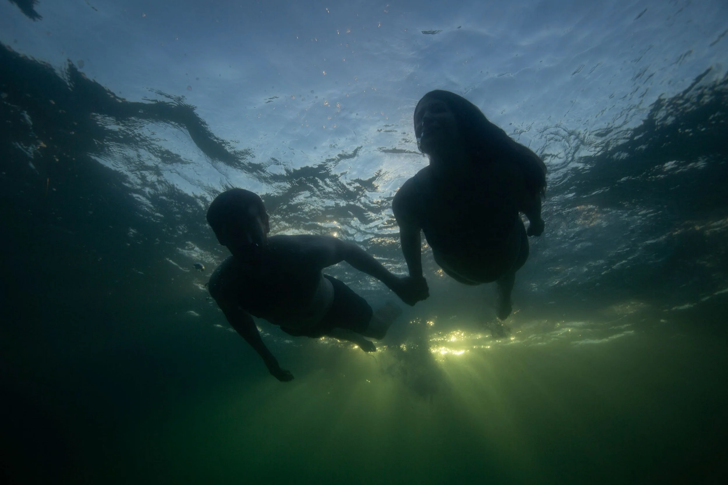 Two people swimming underwater holding hands, with sunlight shining through the water's surface.