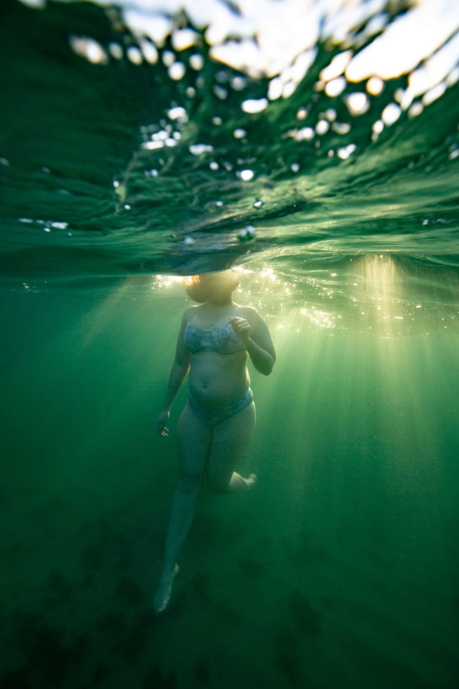 An underwater photograph of a woman in a bikini, with sunlight filtering through the water and creating a shimmering effect.