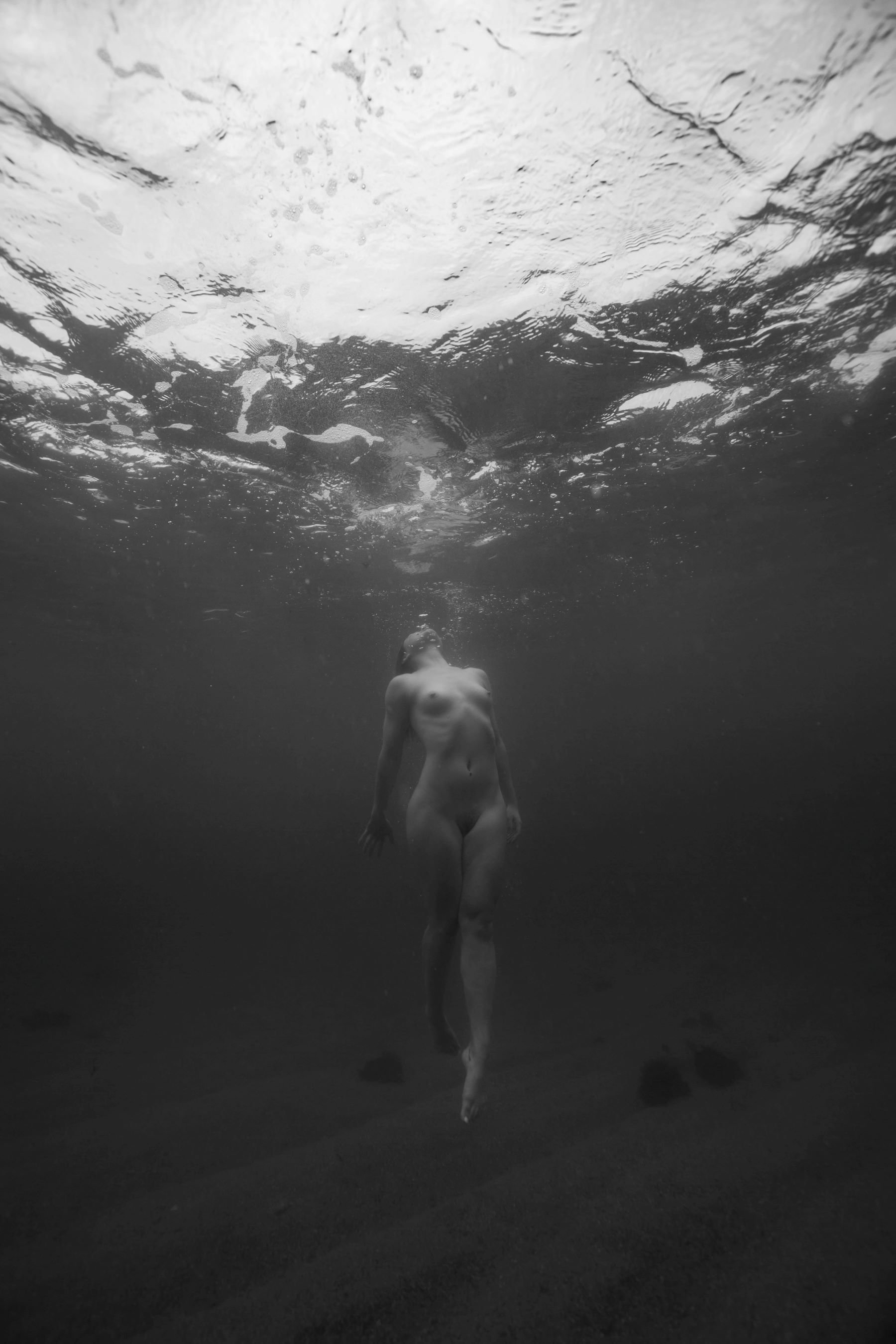 A person swimming underwater with their back to the camera, moving towards the surface, in a black and white photograph.