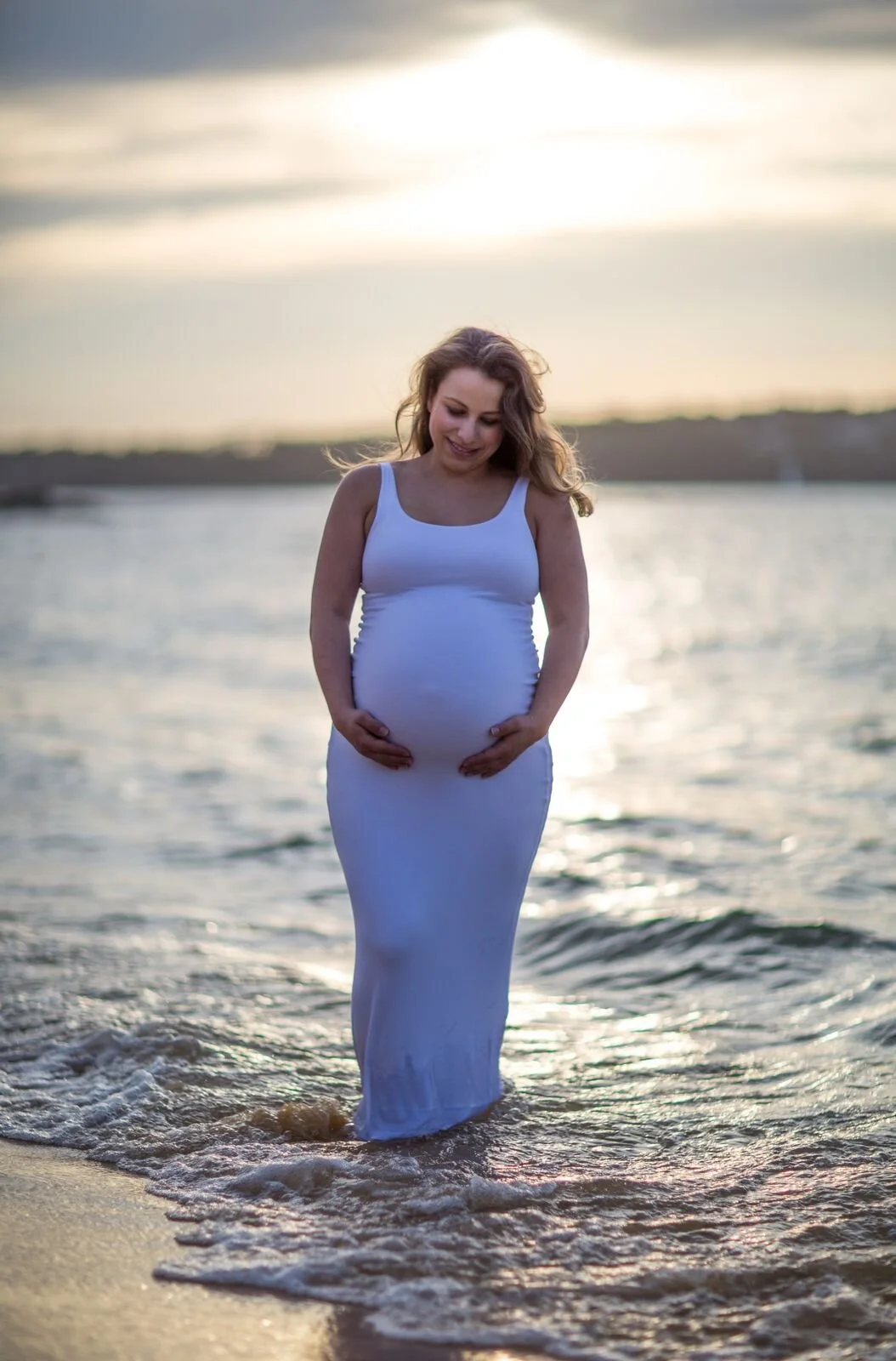 A pregnant woman in a white dress standing in shallow water on the beach during sunset, smiling and holding her belly.  Camp Cove, Sydney.