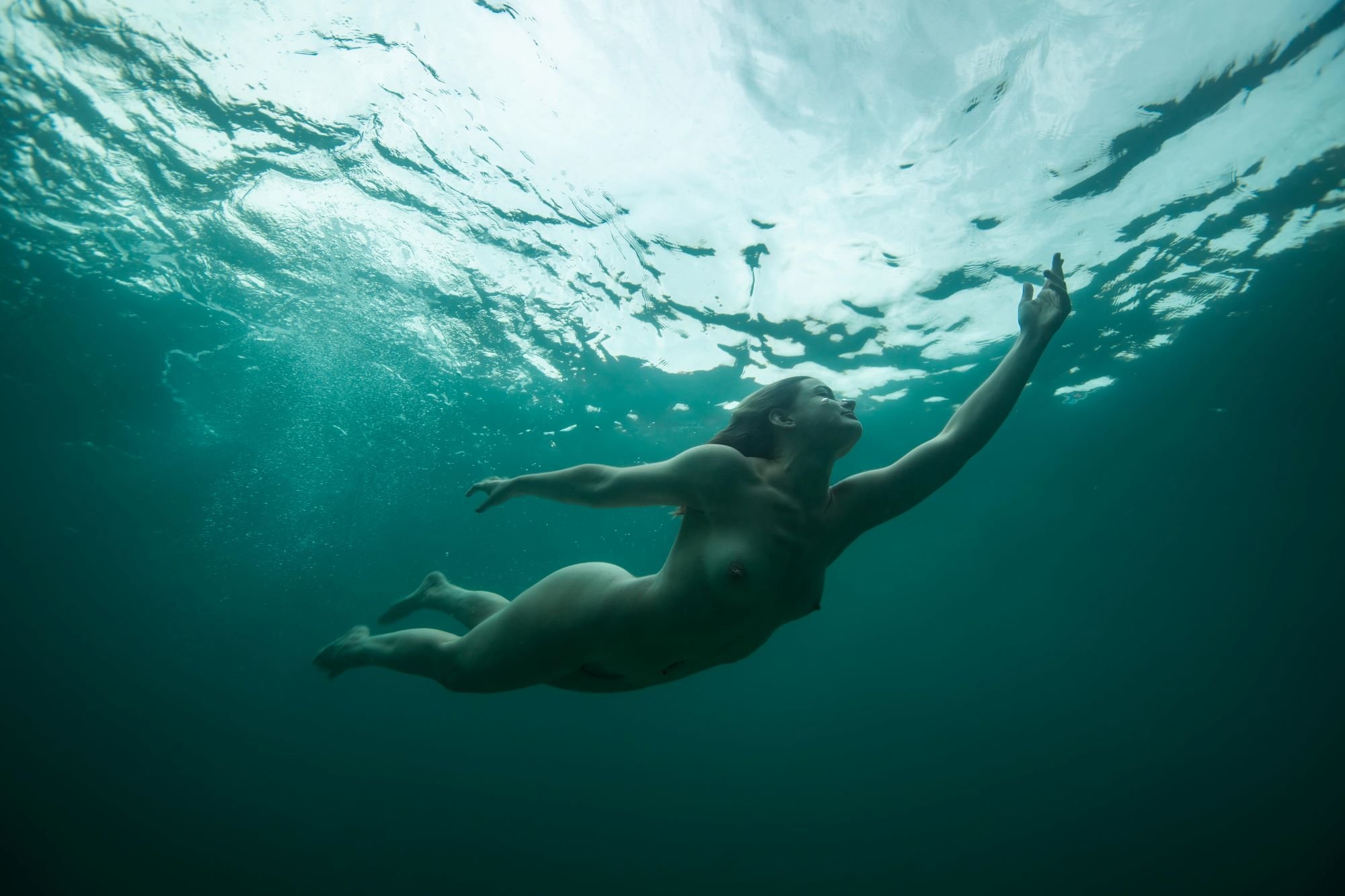 A person swimming underwater, with their body extended horizontally and their head near the surface, reaching upward with one hand. The water is clear with ripples and light filtering from above.