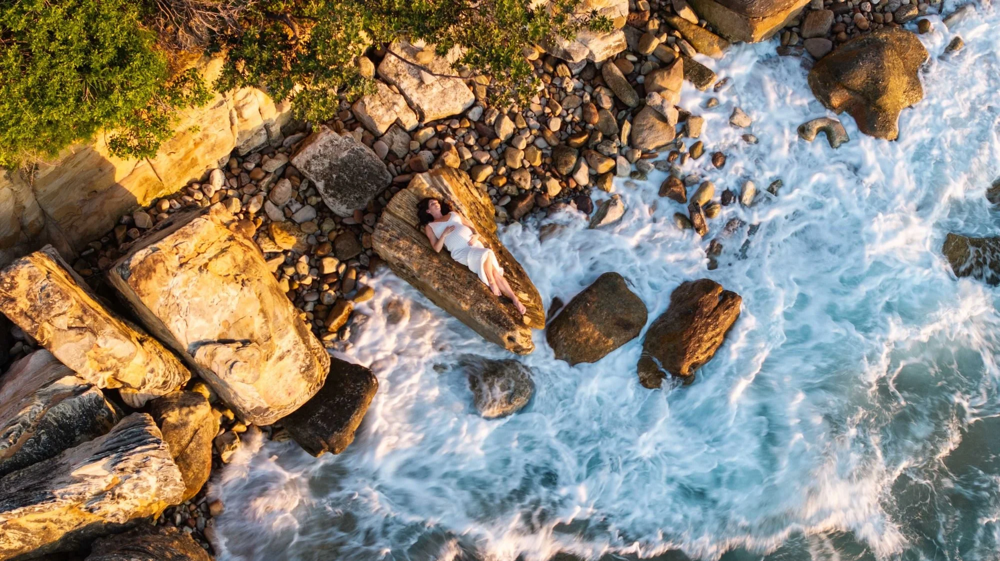 A woman in a white dress lying on a large rock at the edge of the ocean, surrounded by smaller rocks and green trees, with waves crashing nearby during sunset.