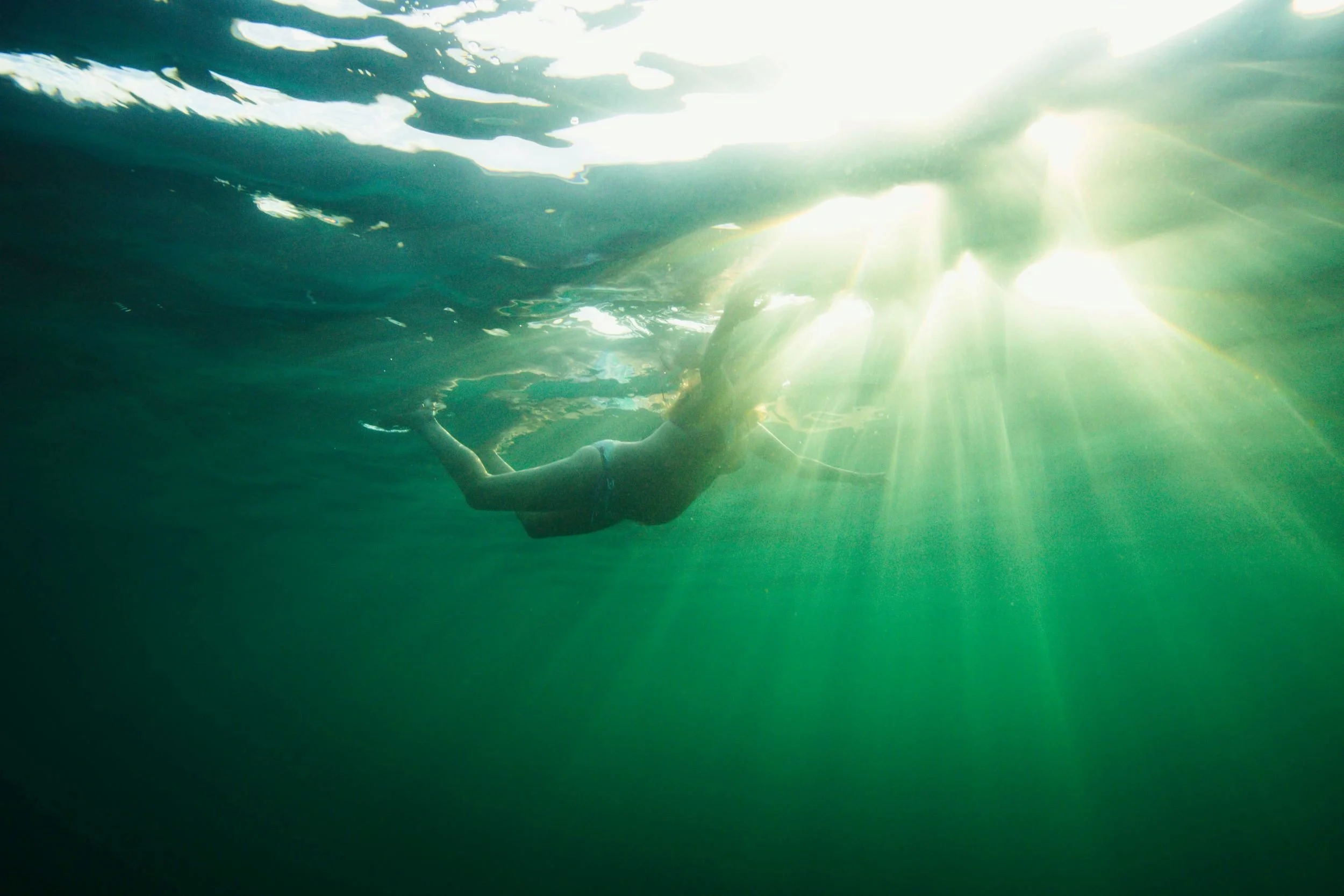 A person swimming underwater near the surface with sunlight streaming through the water.