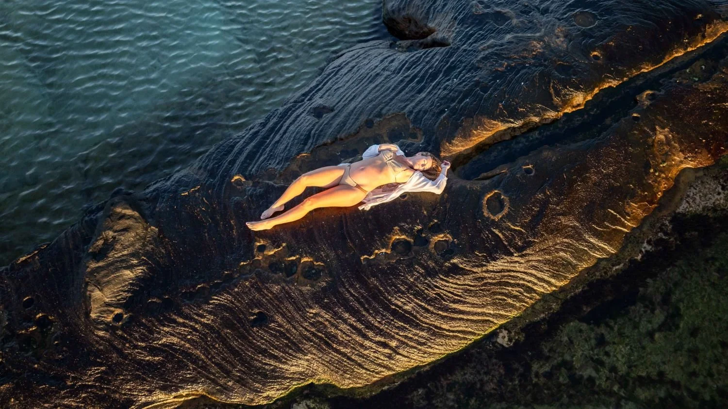 A woman in beige lingerie and a white shirt lying on a large, textured rock near water, during sunset.