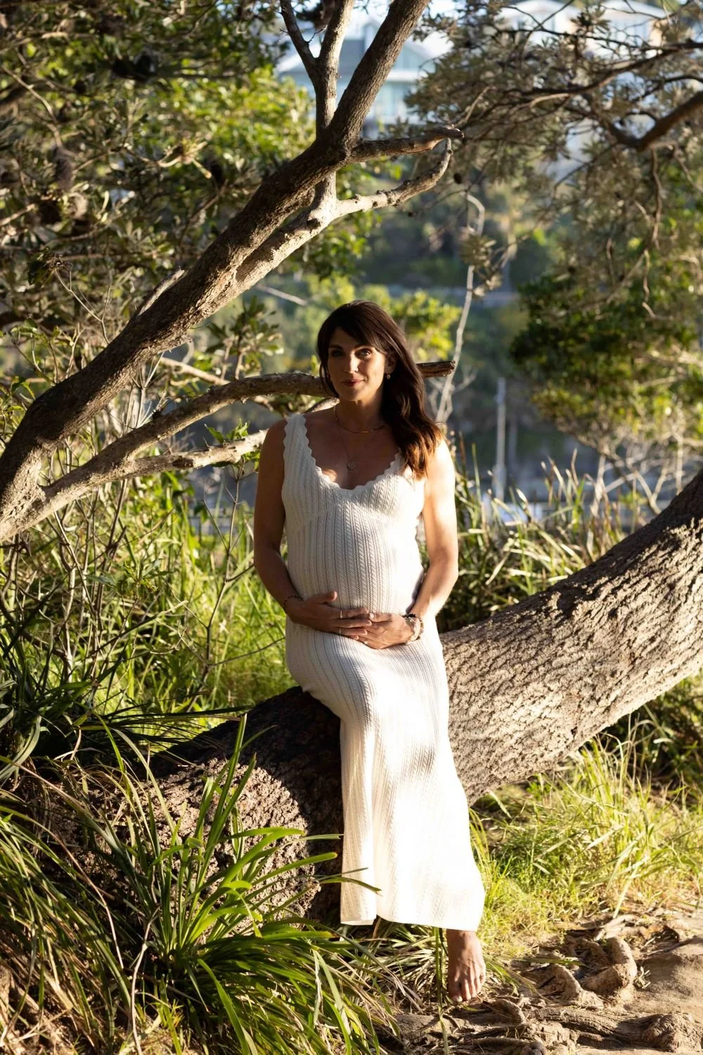 A pregnant woman in a white sleeveless knit dress sitting on a large tree branch in a lush, green outdoor setting during daytime.  Maternity style.