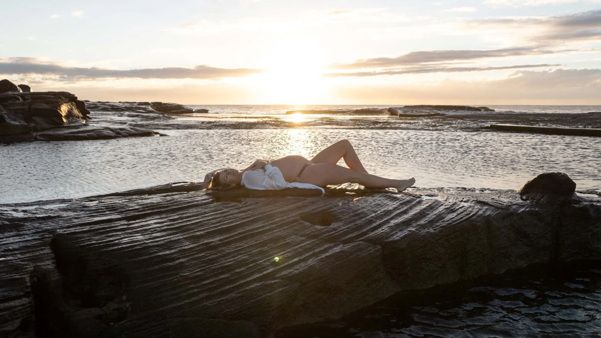 A woman is lying on a rock at the beach during sunset, with water and the ocean in the background and a partly cloudy sky.
