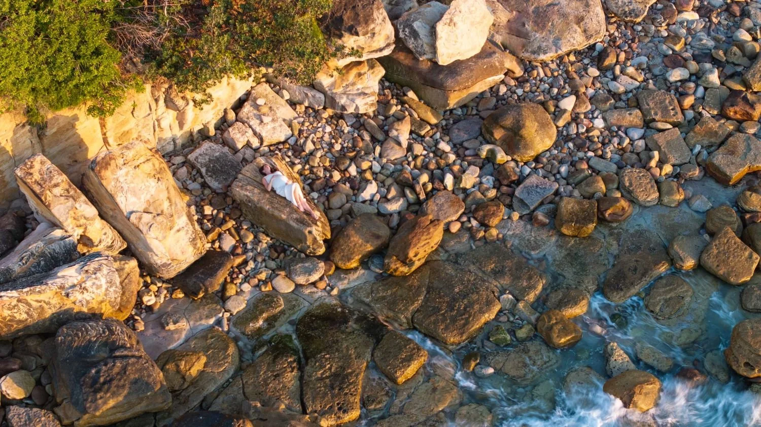 A pregnant lady lying on rocks near the shoreline of a rocky beach, with green bushes above.  Maternity style in Sydney.
