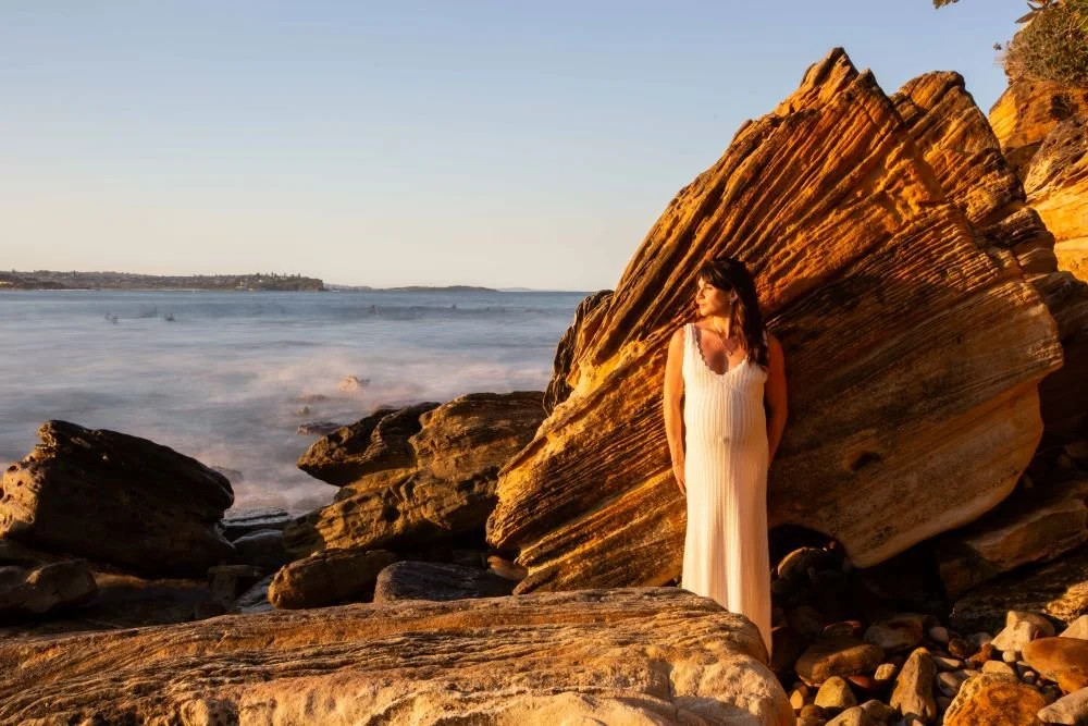 A woman in a white dress standing among large rocks on a beach at sunset, with the ocean and a distant shoreline in the background.