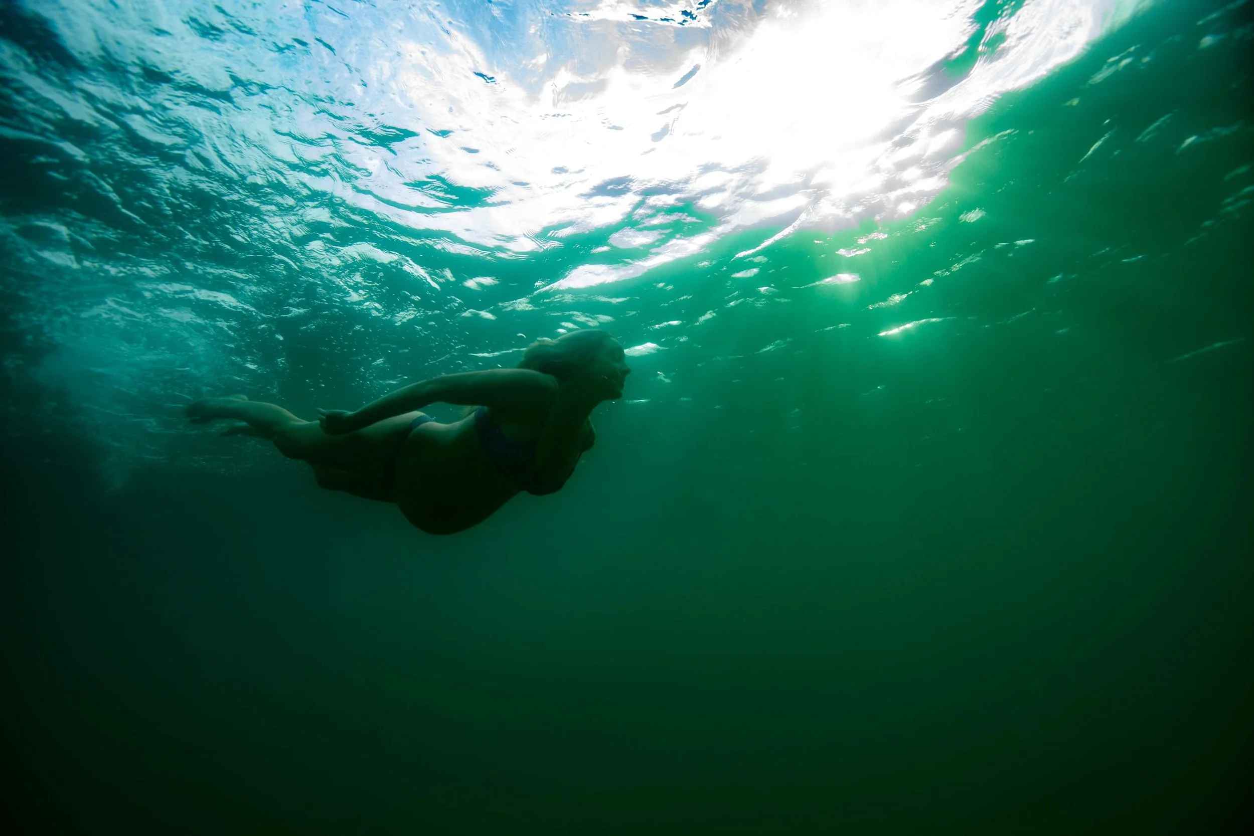 A pregnant woman swimming underwater in the ocean, with sunlight filtering through the water surface and creating a bright, blue-green environment.  Maternity style in Sydney.