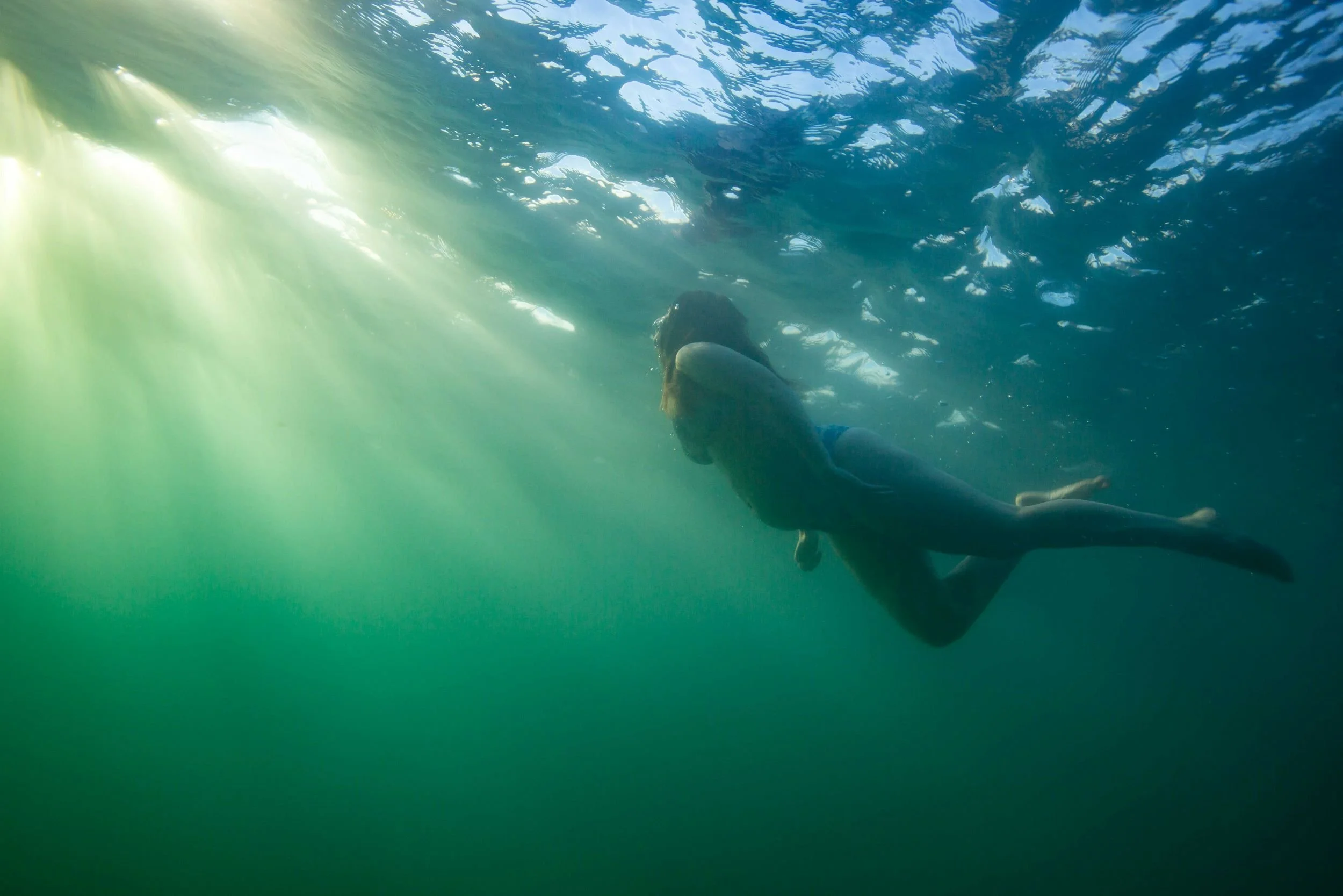 A pregnant person swimming under the water with sunlight filtering through from above.  Maternity style at Manly.