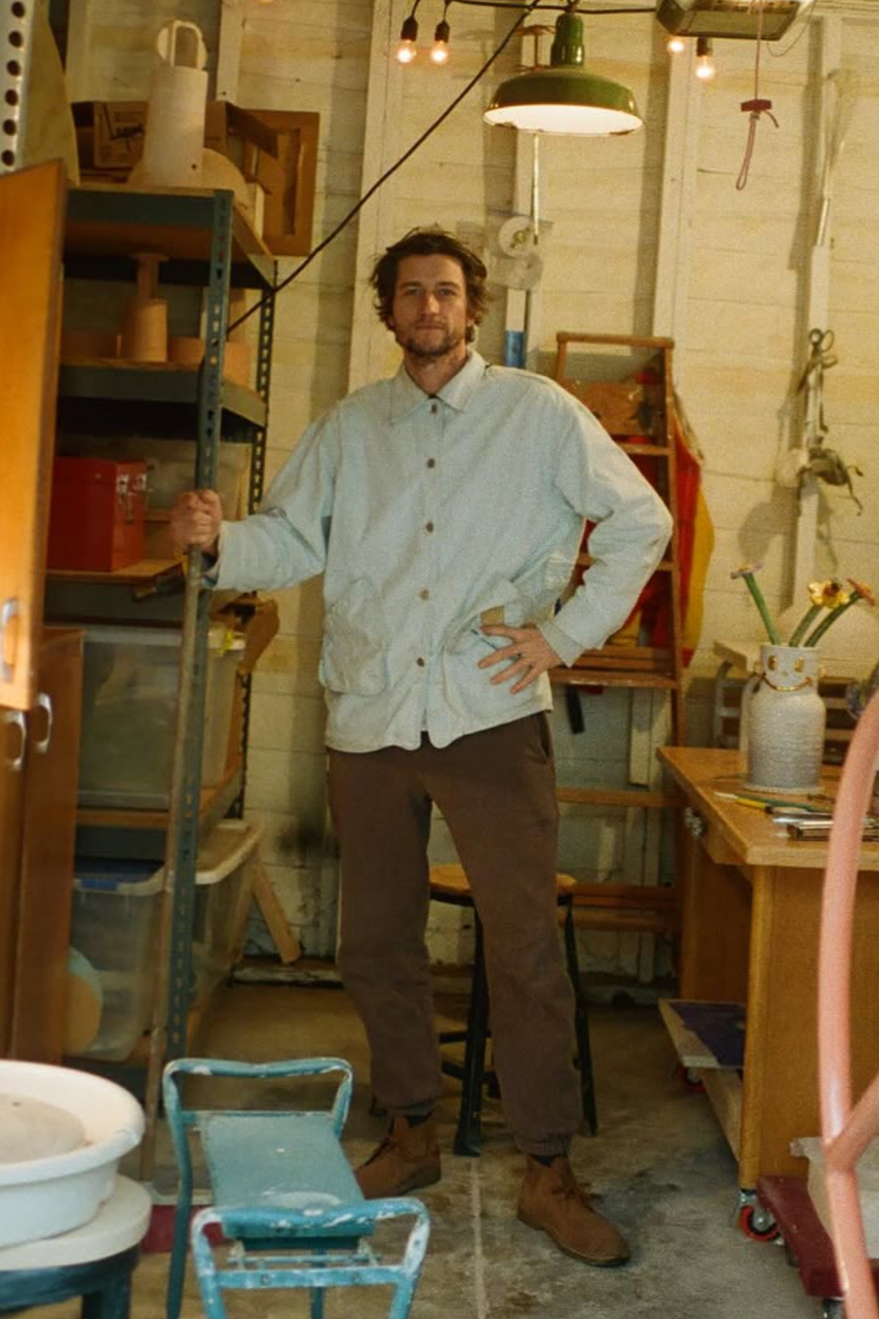 A man standing in a workshop with shelves, a table with a vase and scissors, and various tools and objects around him.