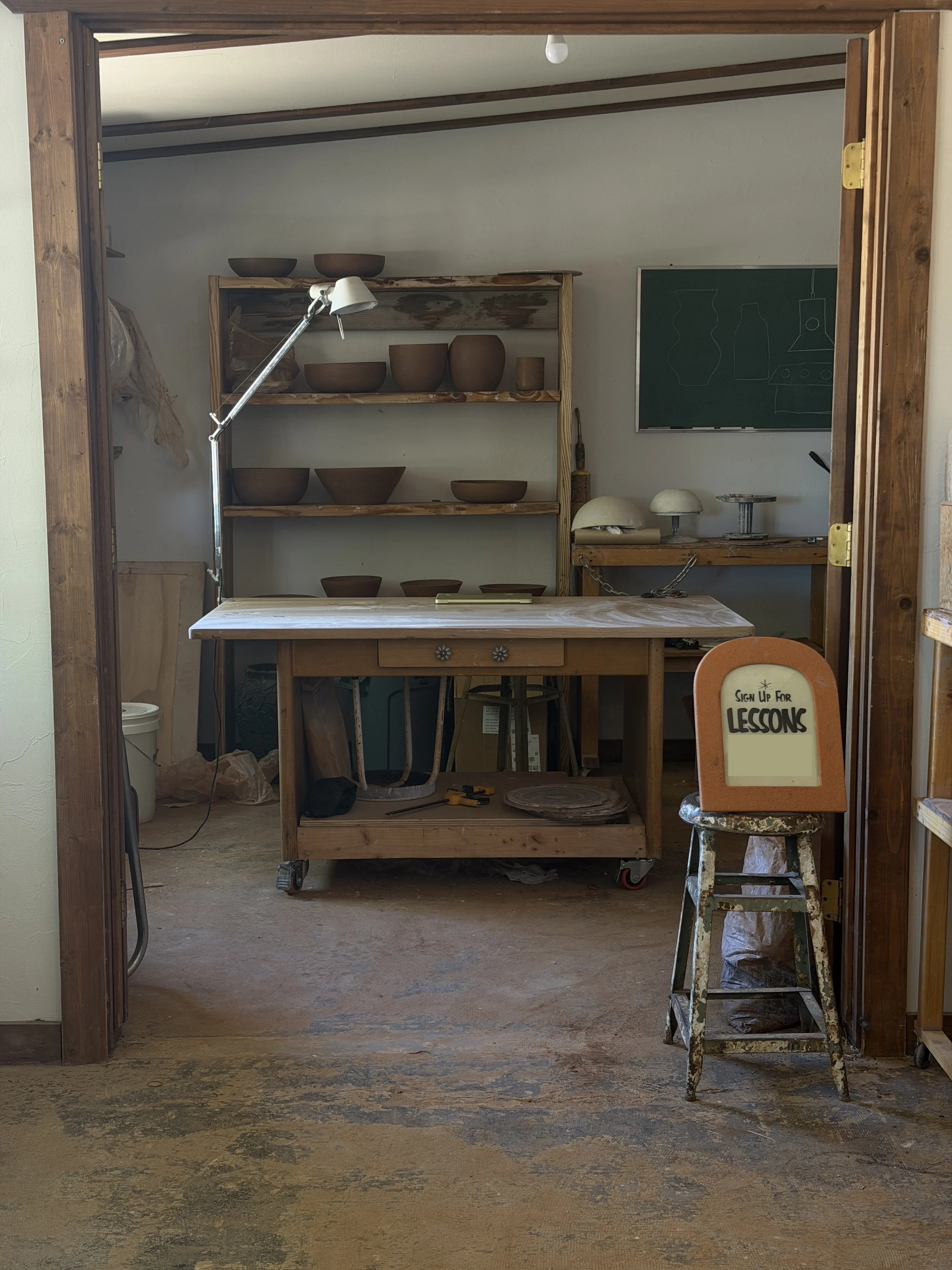 A workshop or classroom with wooden shelving holding pottery bowls and vessels, a blackboard with line drawings of various pottery shapes, a worktable with pottery tools and covered objects, and a vintage metal step ladder with signs of wear and paint splatters, indicating a space used for pottery lessons or studio work.