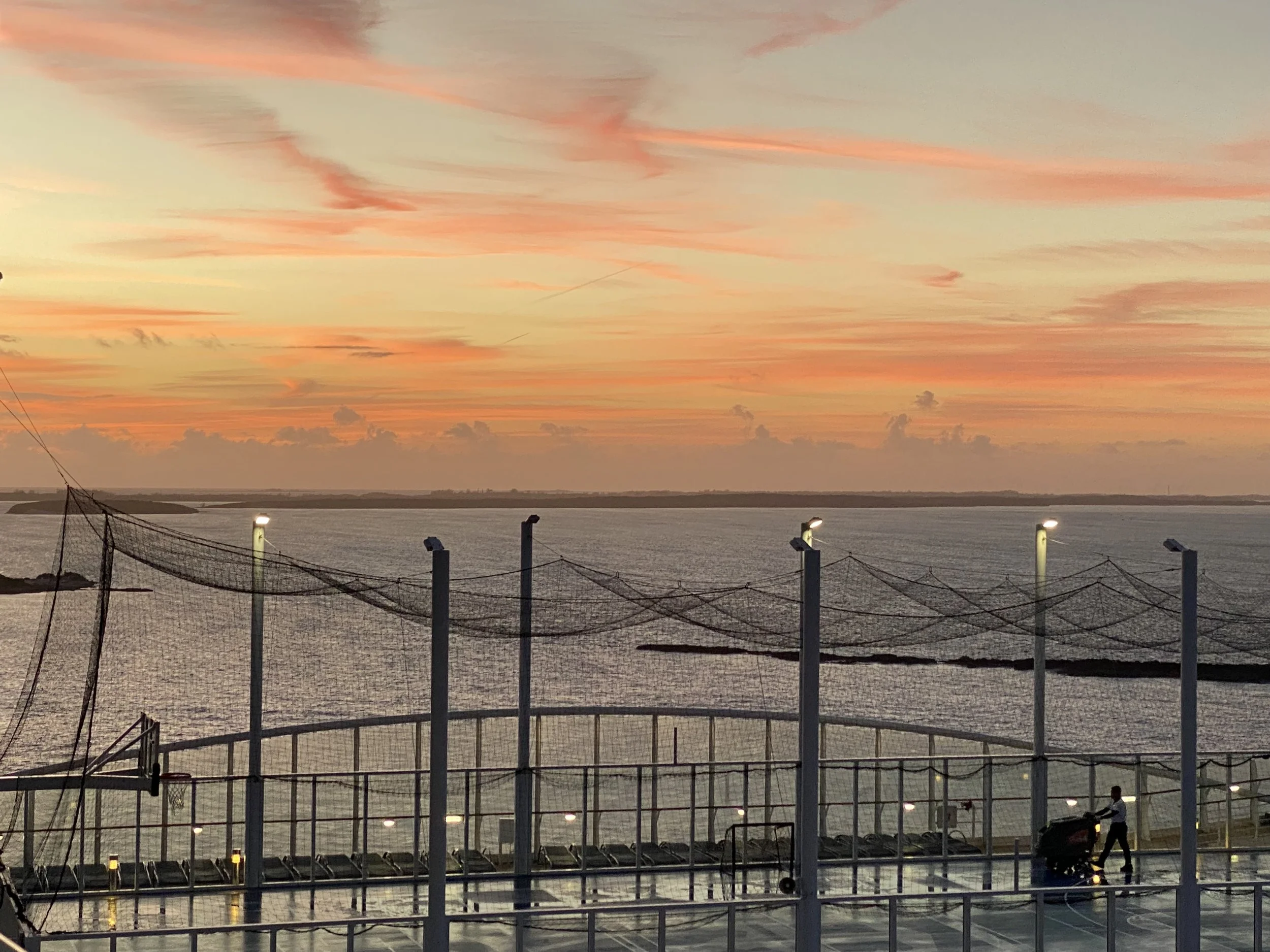 Sunset over a body of water viewed from a sports court with tall poles and nets, and a person pushing a cart.