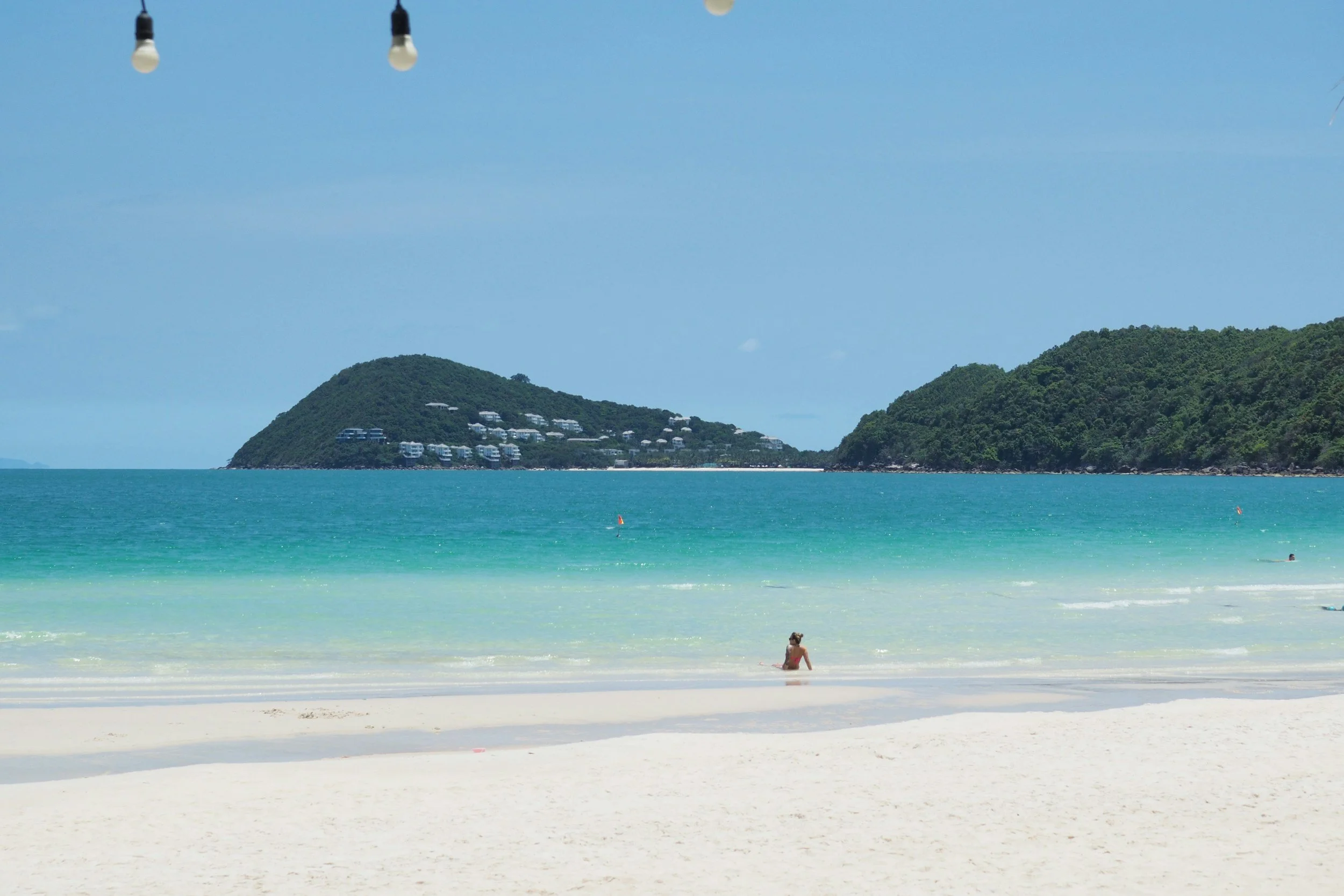 Sandy beach with a person sitting by the shore, turquoise ocean water, green forested hills in the distance, clear blue sky, and string lights hanging at the top of the image.