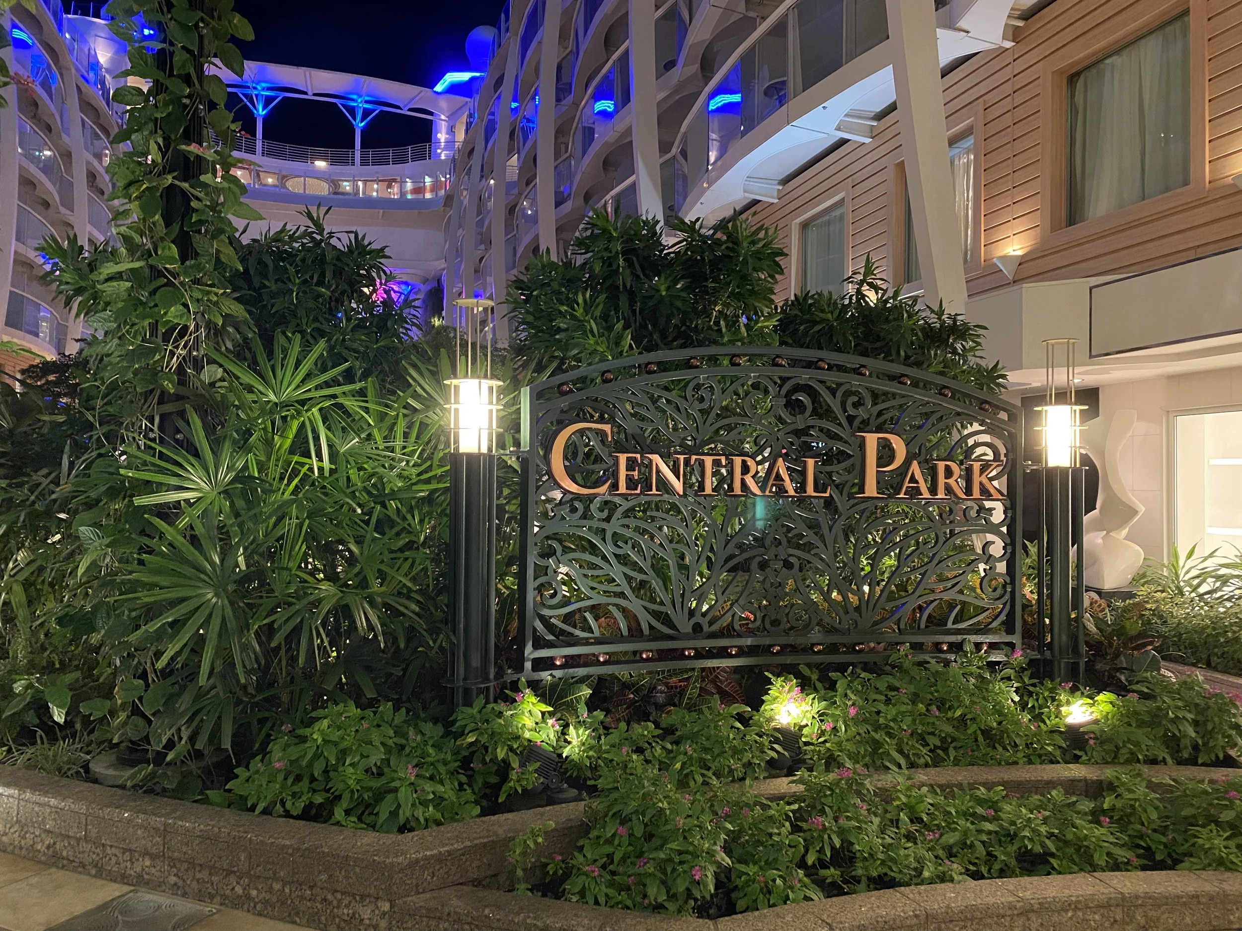 Night view of the Central Park sign on a decorative wrought iron gate, surrounded by lush greenery, with the building's exterior and balconies lit by blue, white, and warm lighting.