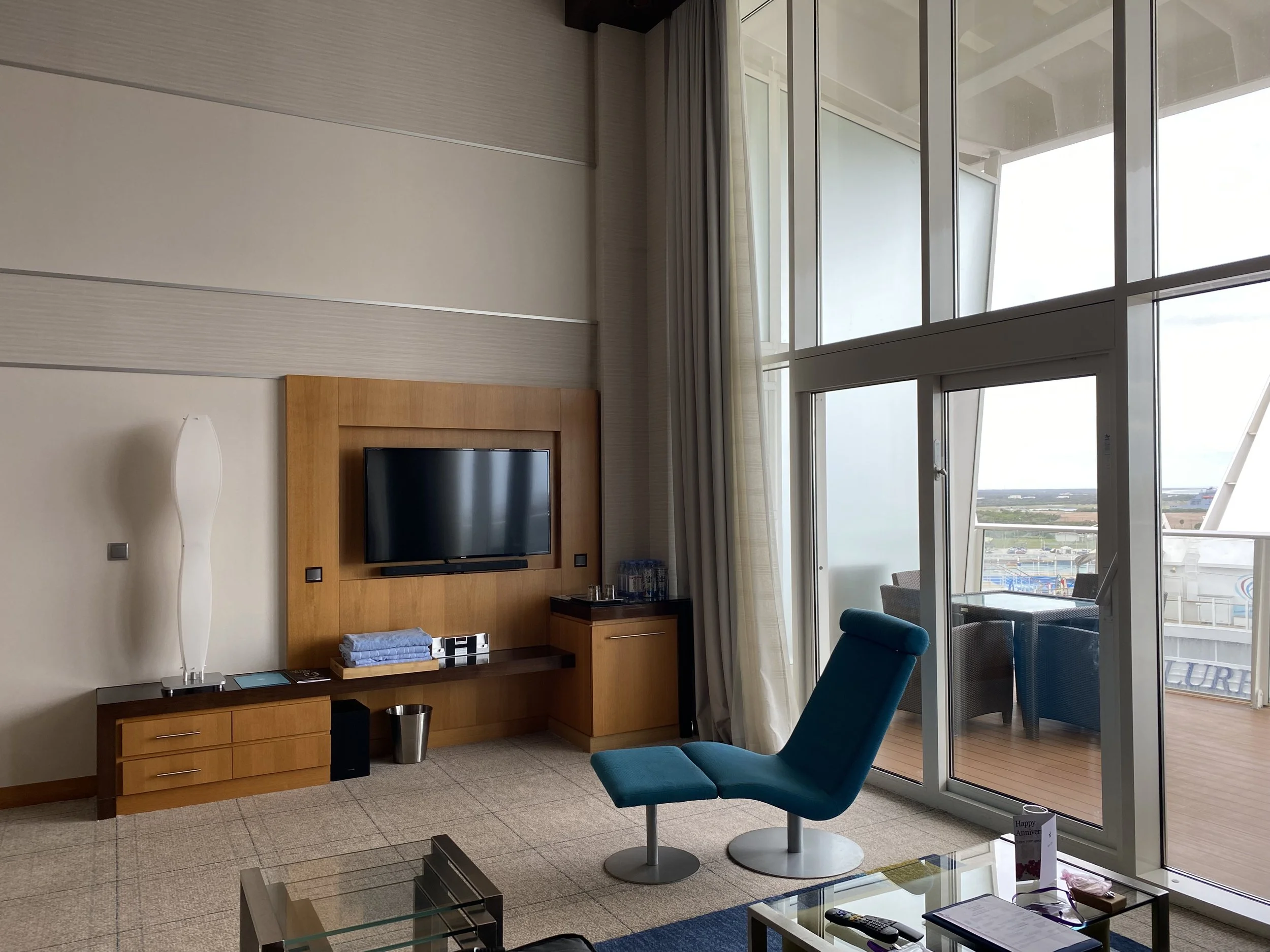 A hotel room with a blue reclining chair, a wall-mounted flat-screen TV on a wooden panel, a side table with water bottles, and a balcony with outdoor furniture visible through large glass sliding doors.