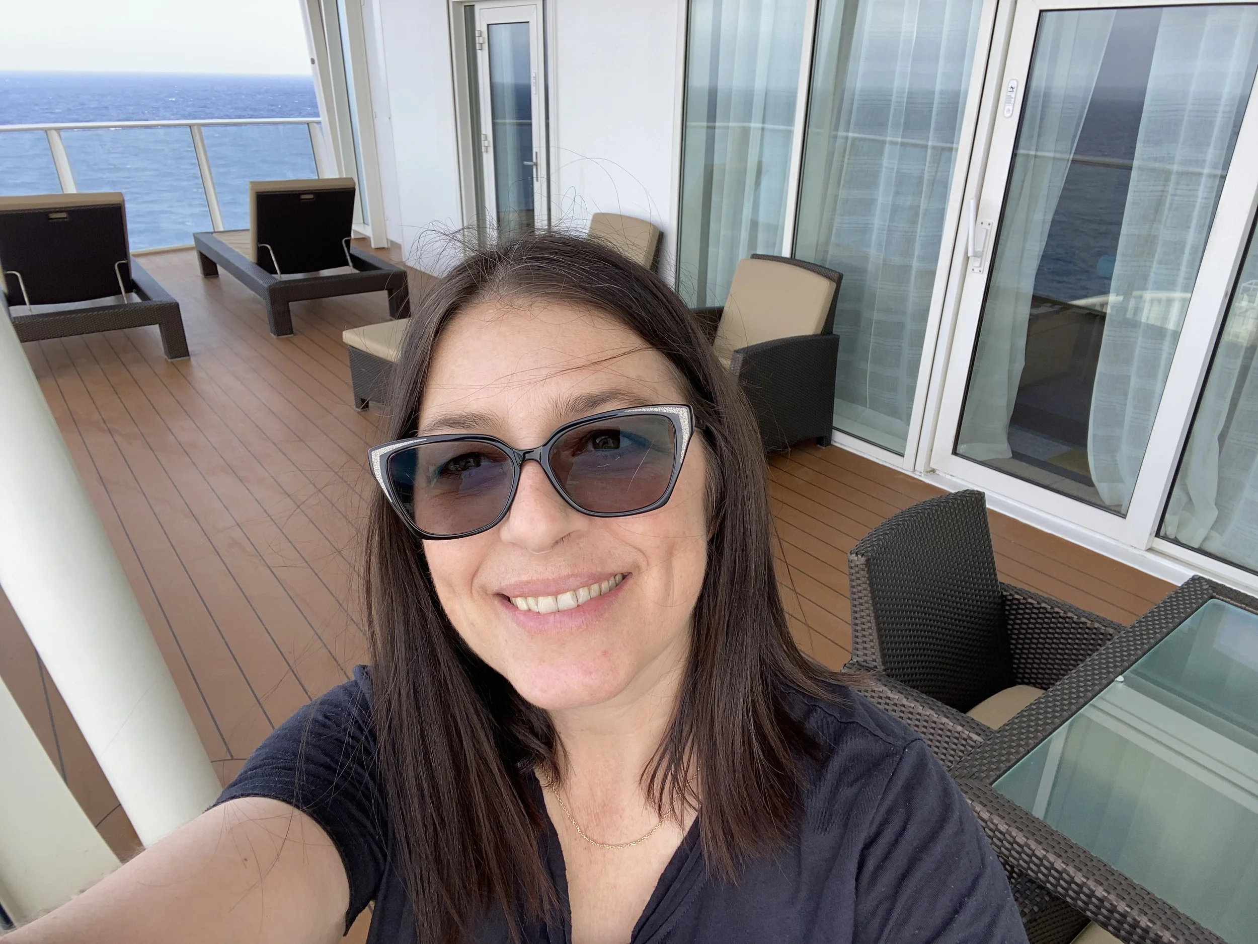 Smiling woman wearing sunglasses on a cruise ship balcony with ocean view, patio furniture, and a sliding glass door.