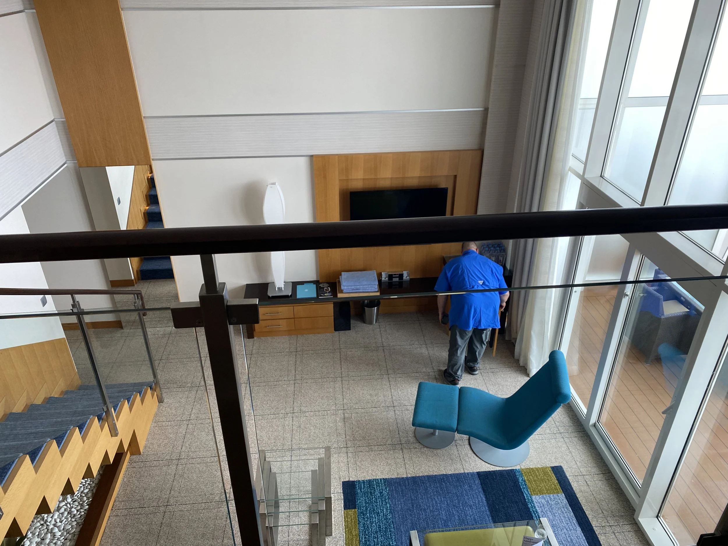 Interior of a hotel lobby viewed from an elevated position, featuring a man in a blue jacket cleaning near a large window, a teal lounge chair, a colorful area rug, and stairs leading to an upper level.