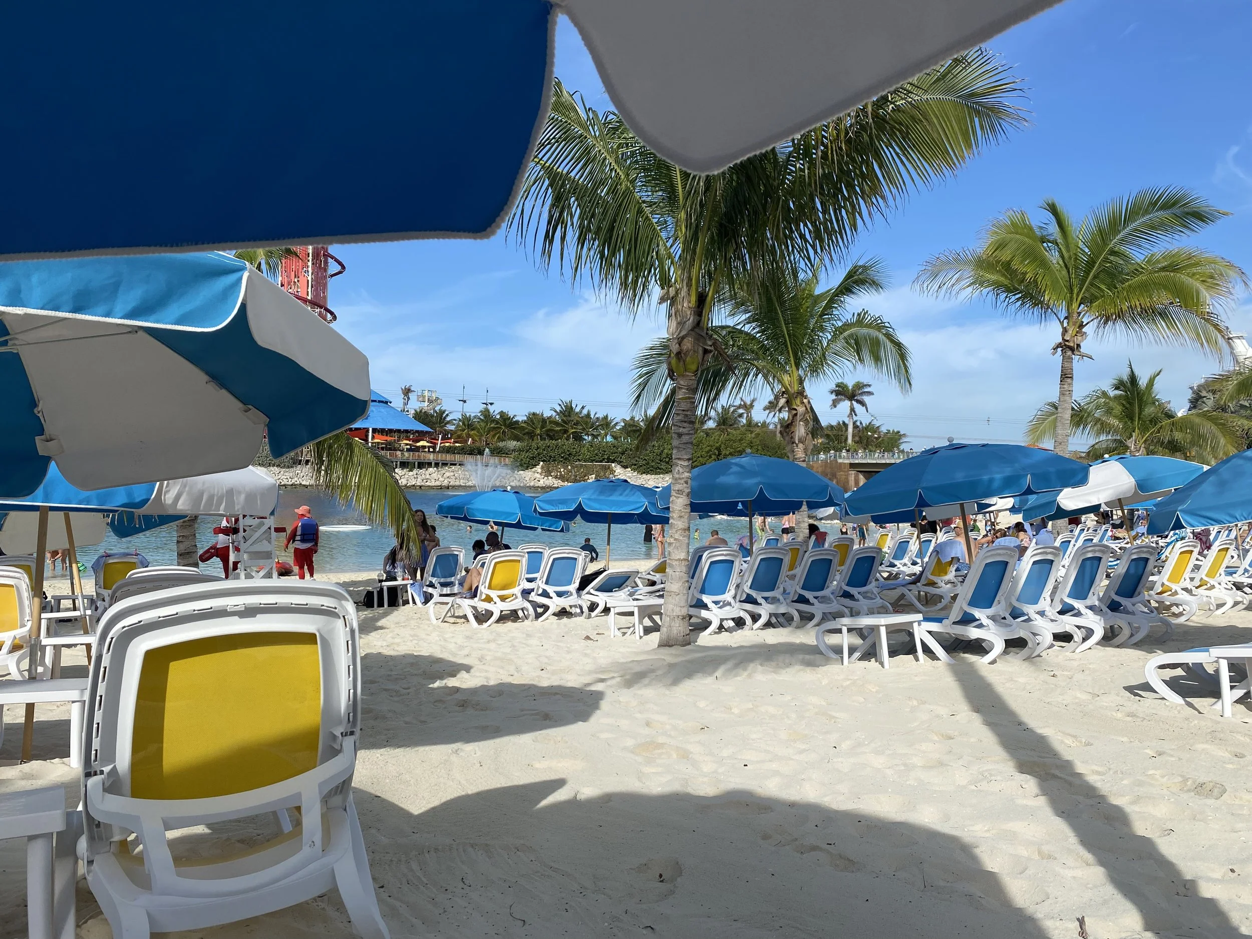 Beach with white sand, blue umbrellas, and white lounge chairs. Palm trees line the background, and a water feature is visible near the shore.