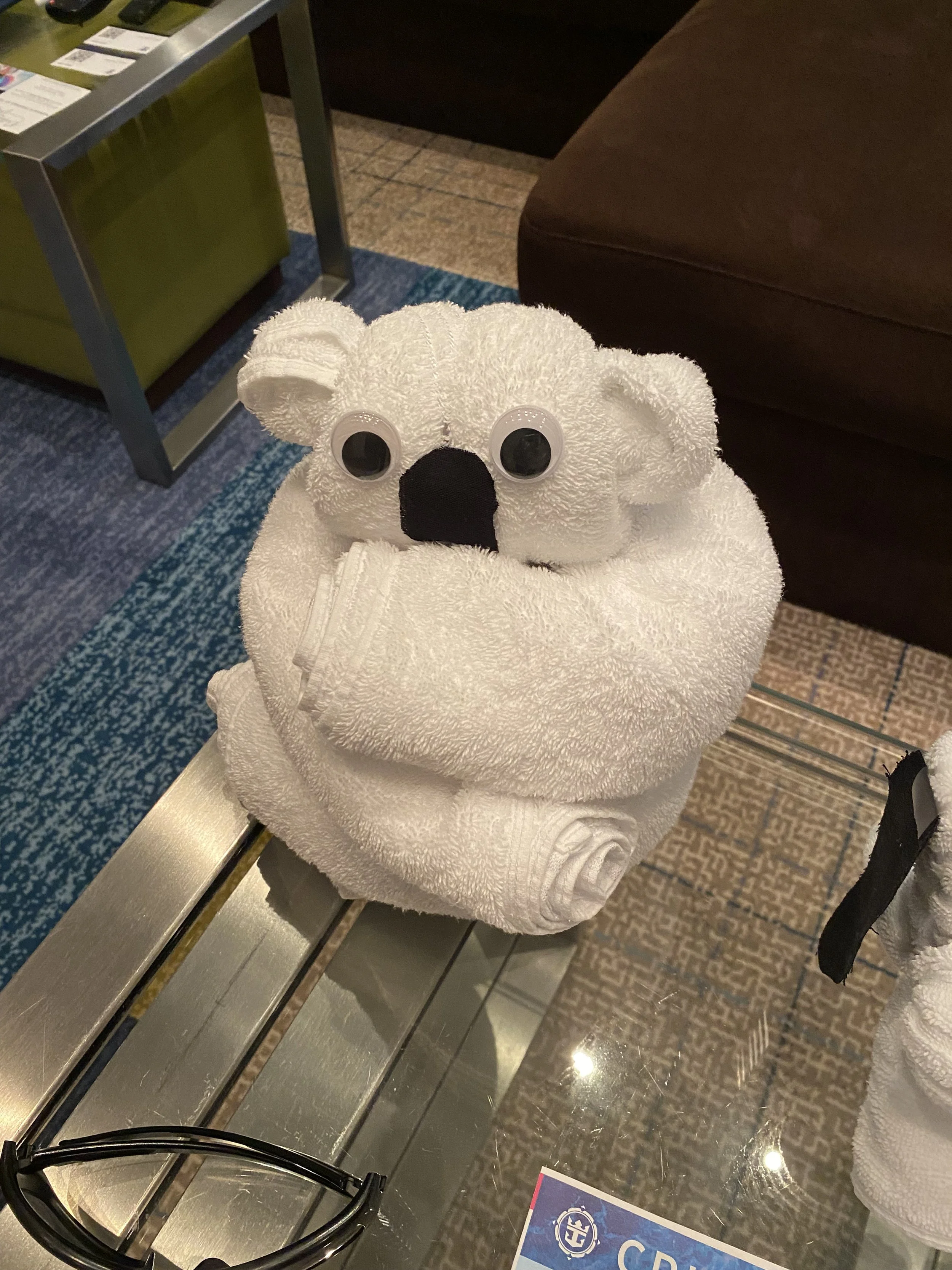 A stuffed koala bear made from a white towel with googly eyes and a black nose, sitting on a glass surface in a hotel lobby or retail store.