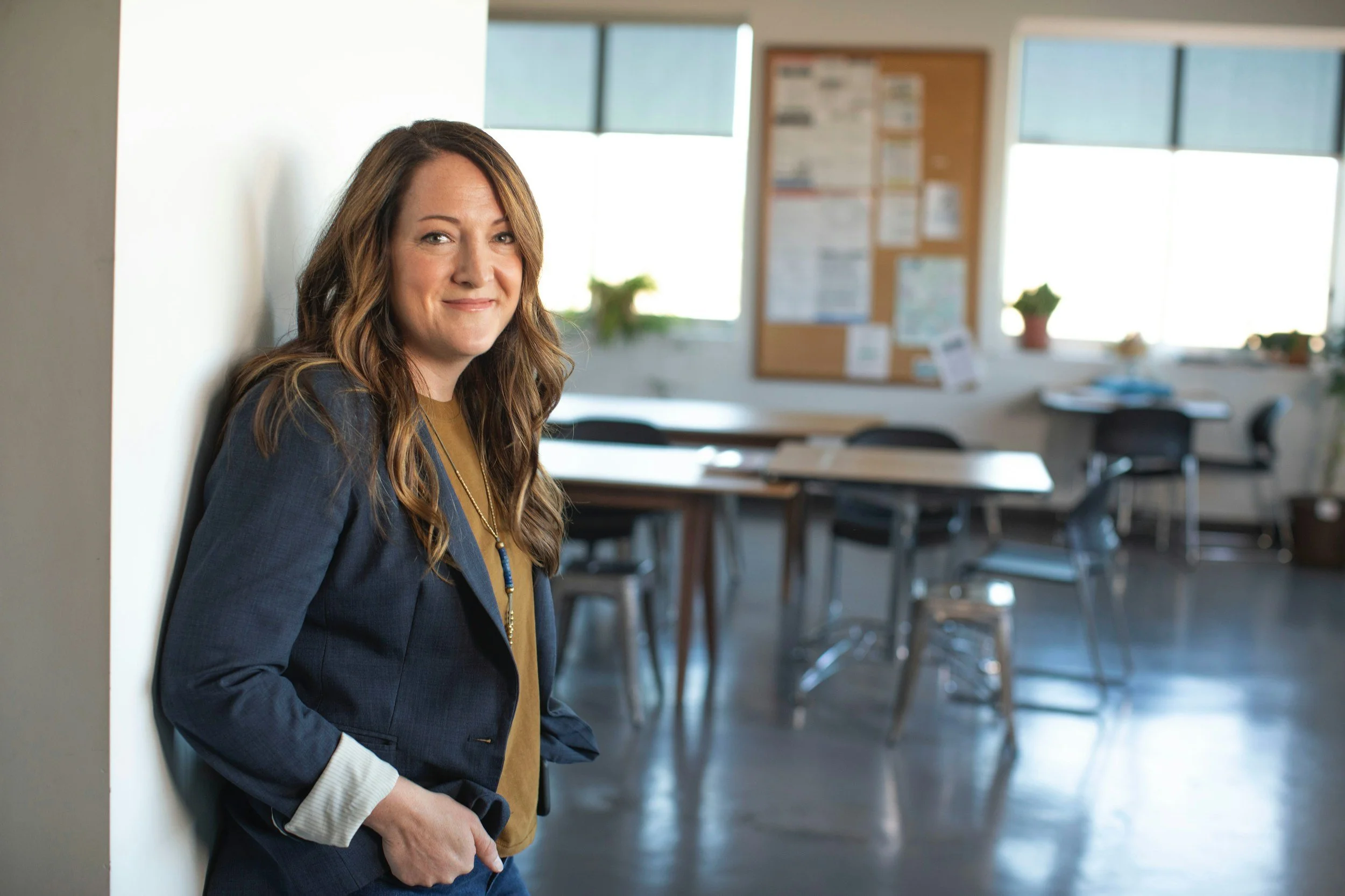 A woman with long brown hair wearing a navy blazer and tan top, standing in a classroom or office space and leaning against a wall, smiling at the camera.