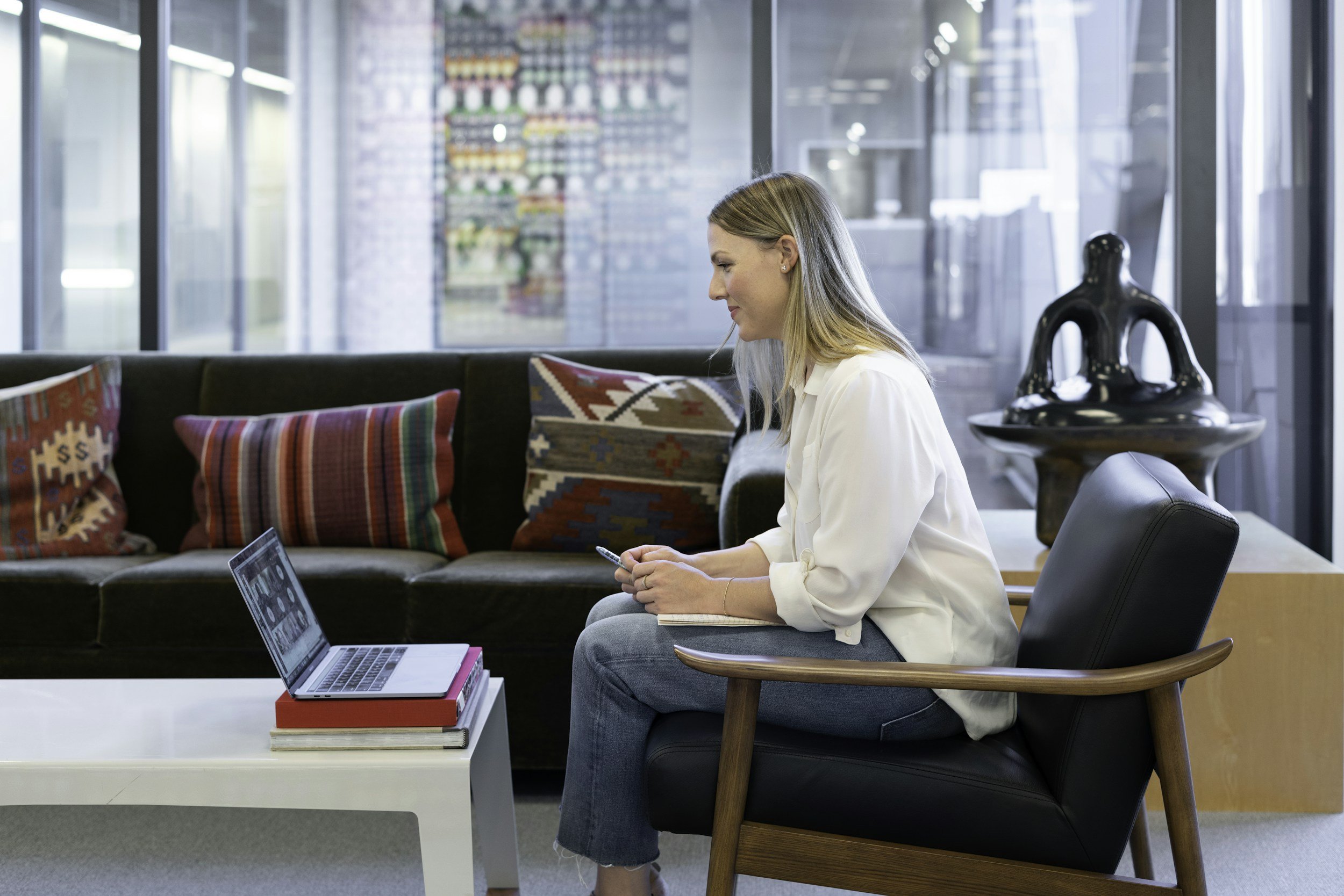 A young woman with blonde hair sitting on a black chair in a modern office or lobby area, using her phone. She is wearing a white blouse. There are four colorful patterned pillows on a black sofa behind her. In front of her is a white table with a stack of books and an open laptop. The background features large glass windows and a decorative sculpture, with a blurred colorful wall art visible through the window.