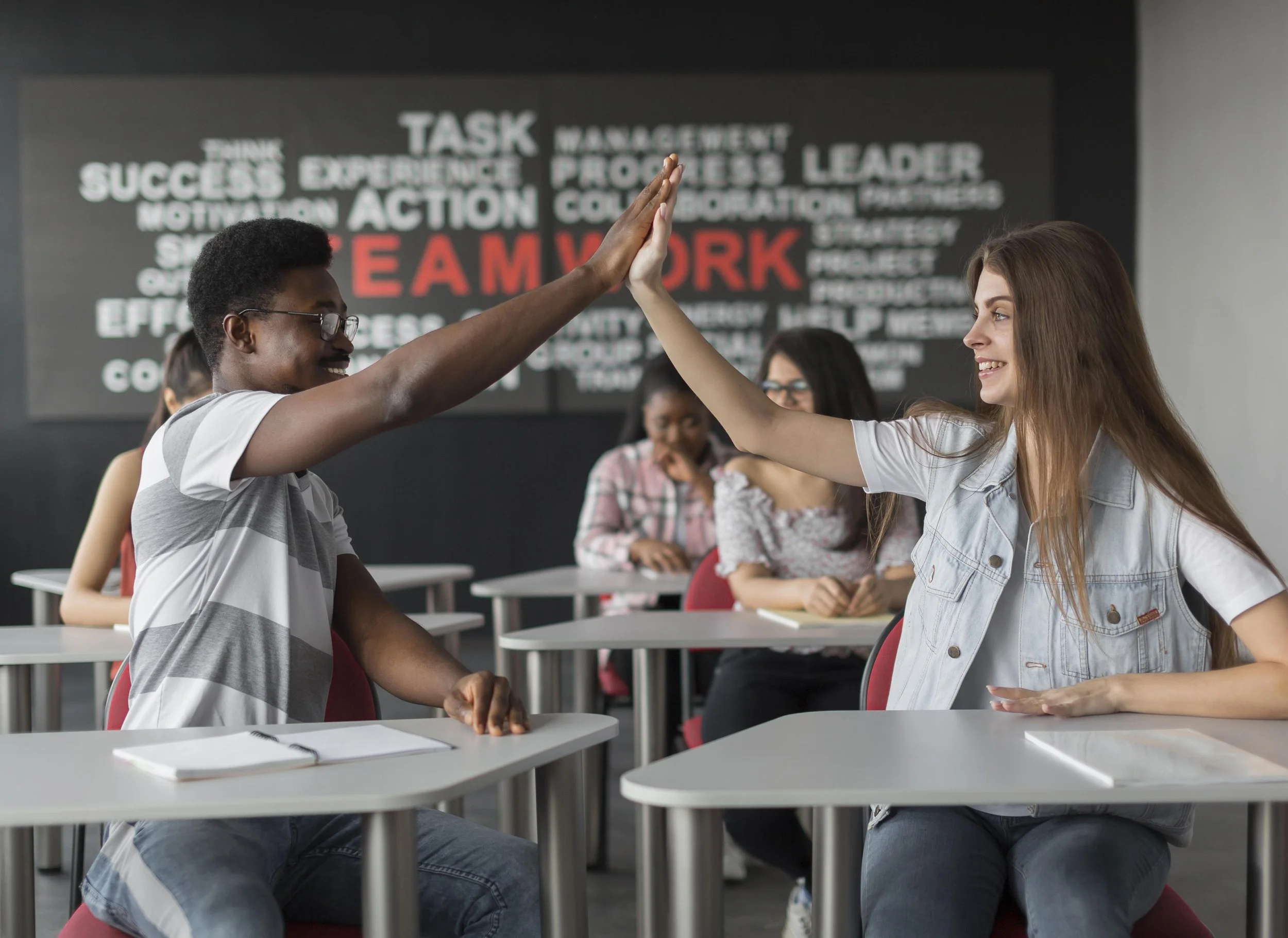 Two women, one with dark skin and glasses and the other with light skin and long brown hair, are giving each other a high-five in a classroom setting. Other students are visible in the background, and a large word wall with the word 'TEAMWORK' in red is on the wall.