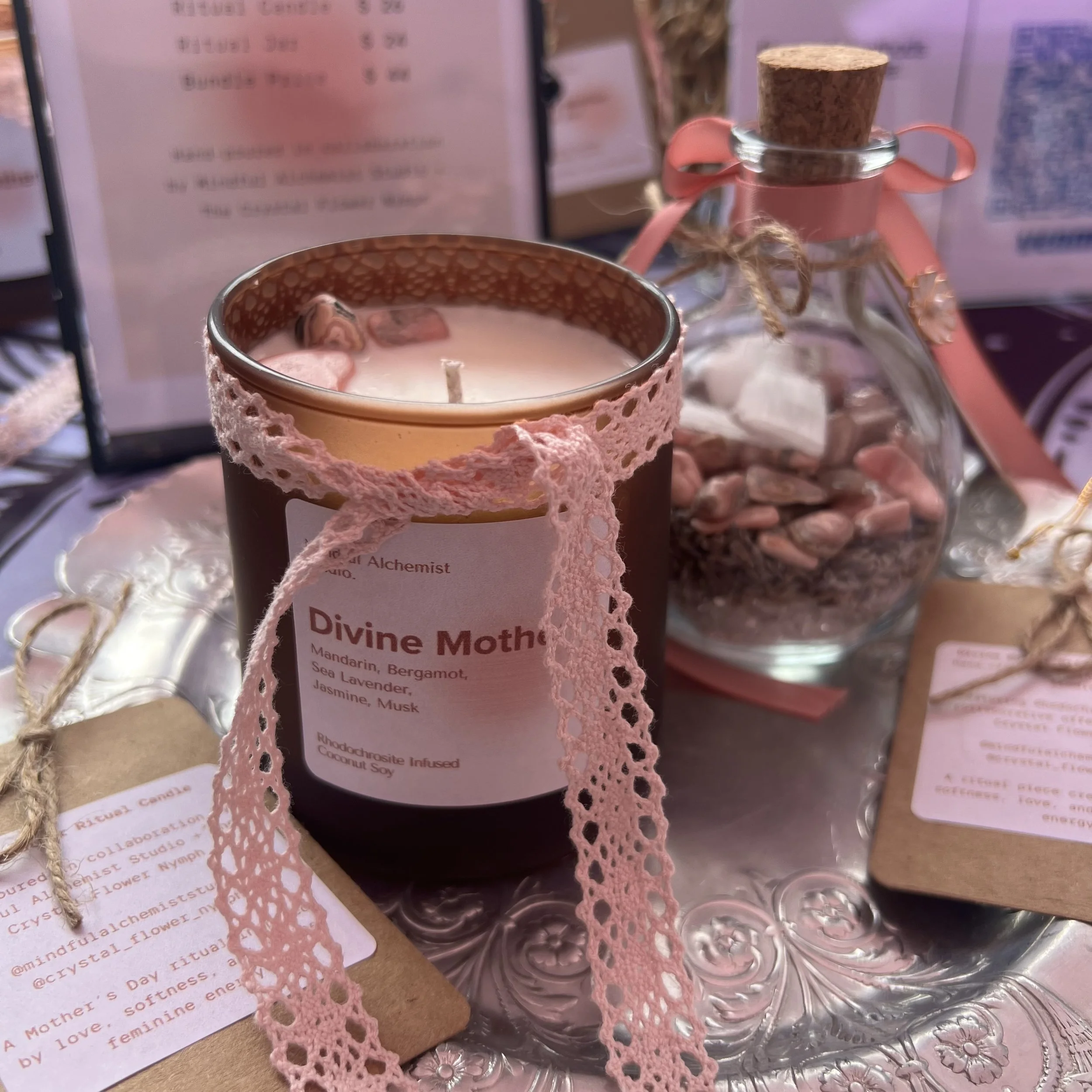 A pink lace-wrapped candle labeled 'Divine Mother' with a glass bottle tied with pink ribbon behind it on a vintage metal tray.