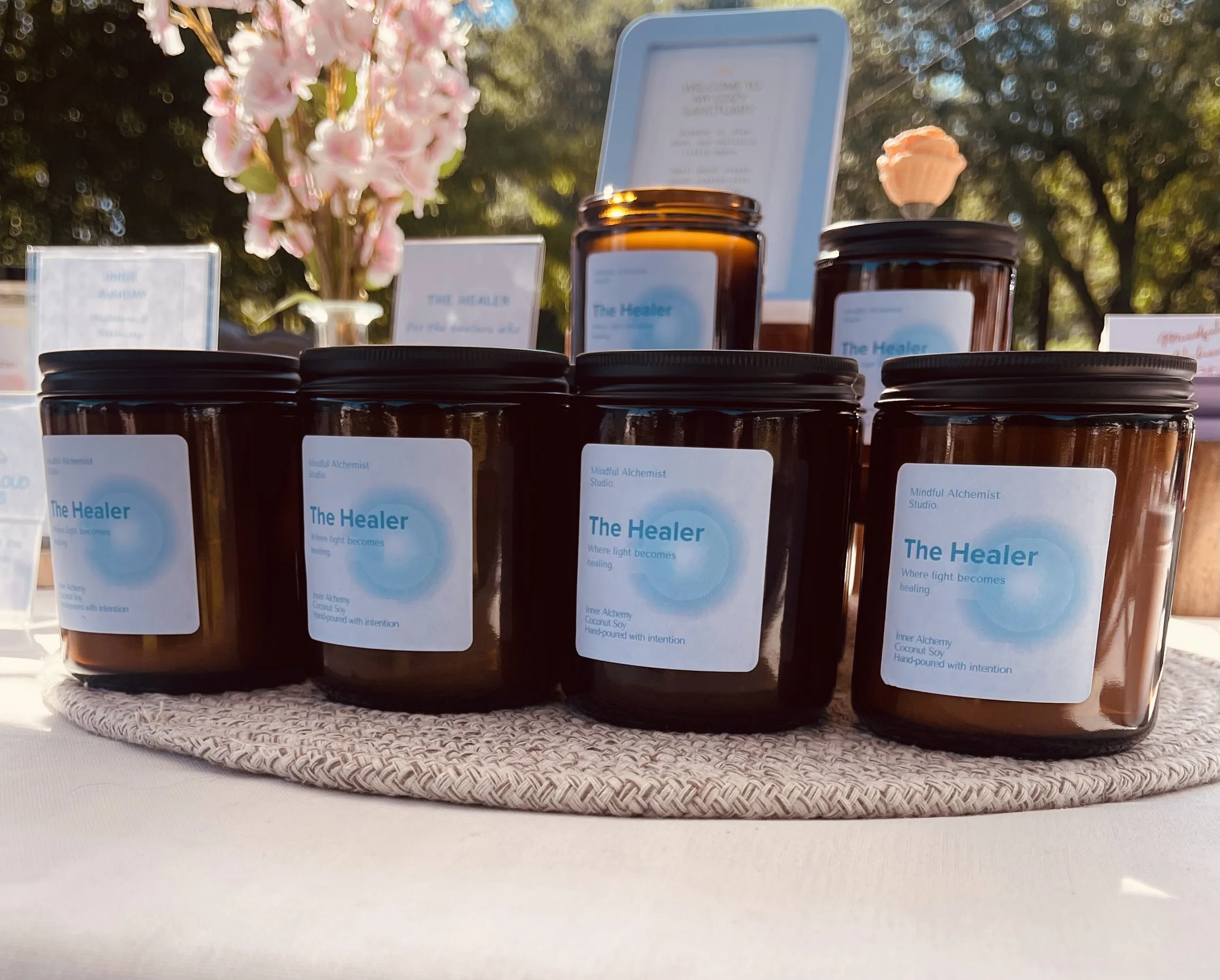 Several amber glass jars with black lids on a woven mat, with labels reading "The Healer" and containing products from a studio called 'Mindful Alchemist Studio.' A pink flower arrangement and a glass display with text are in the background, outdoors with trees and sunlight.
