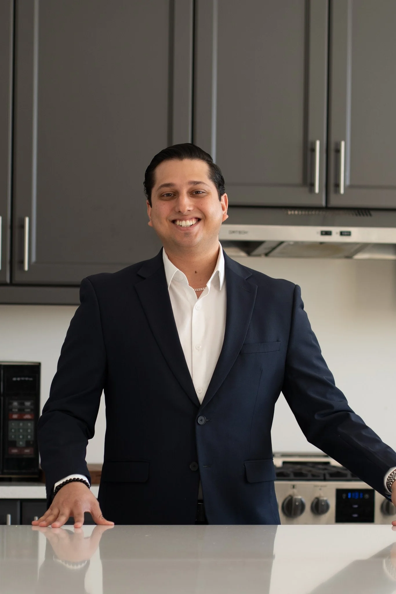 Smiling man in a dark suit standing in a modern kitchen with gray cabinets, white walls, and stainless steel appliances.