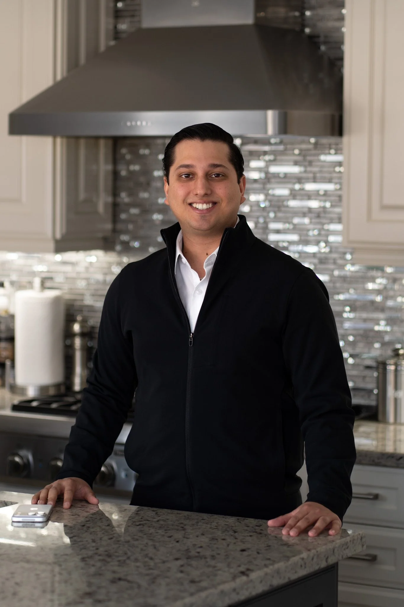 A man smiling while standing in a modern kitchen with a black jacket and white shirt, on a granite countertop.