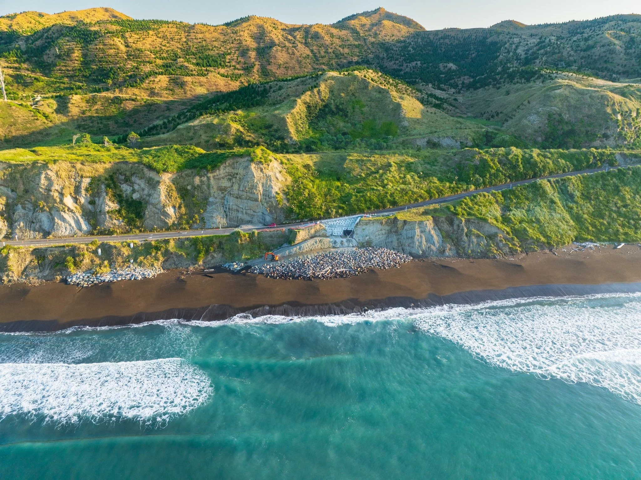 Aerial view of a coastal highway along a cliffside with ocean waves hitting the shore, lush green hills behind the highway, and construction equipment on the rocky shoreline.
