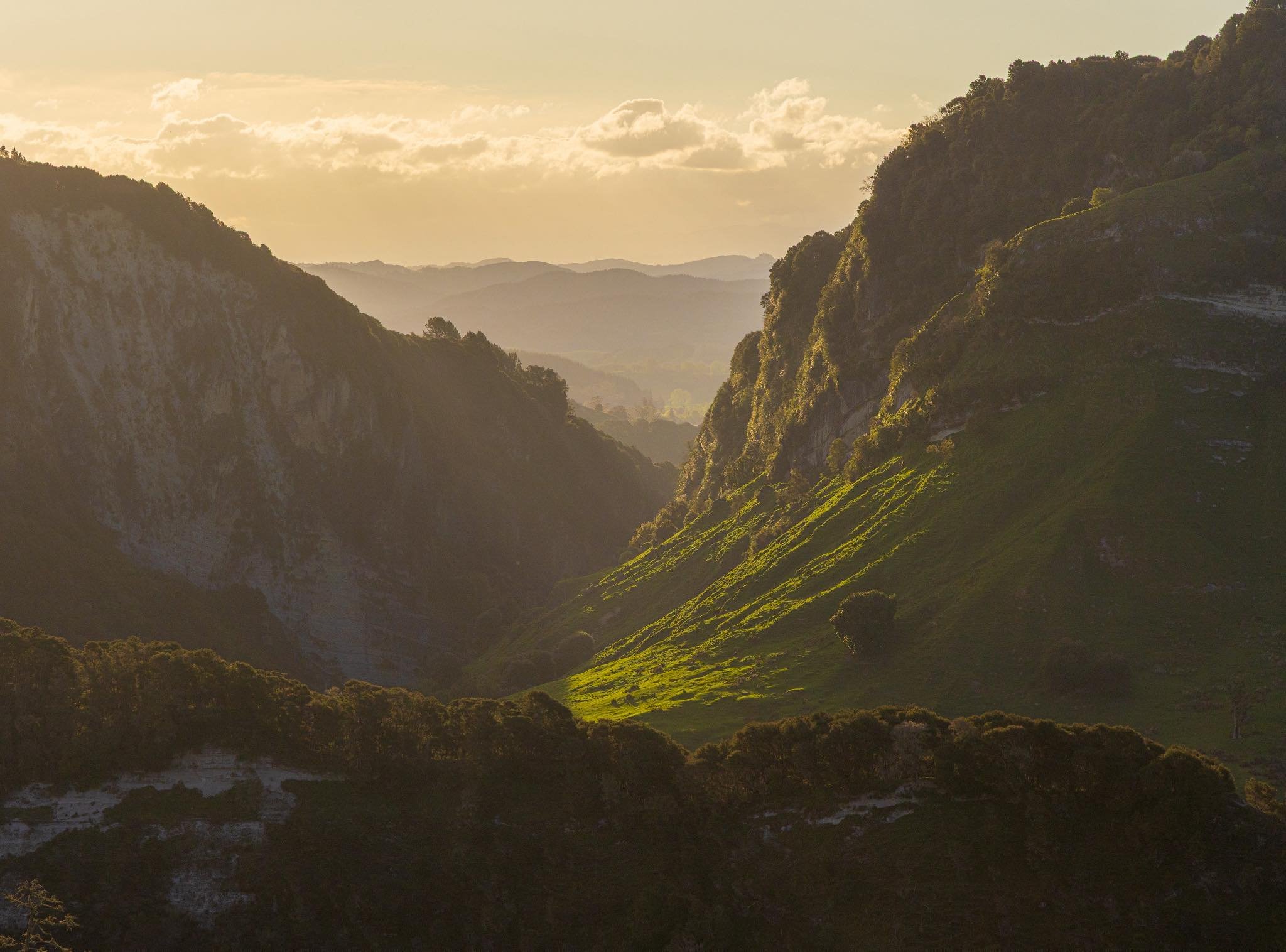 Sunlit green mountains and valley with trees and rolling hills under a partly cloudy sky.