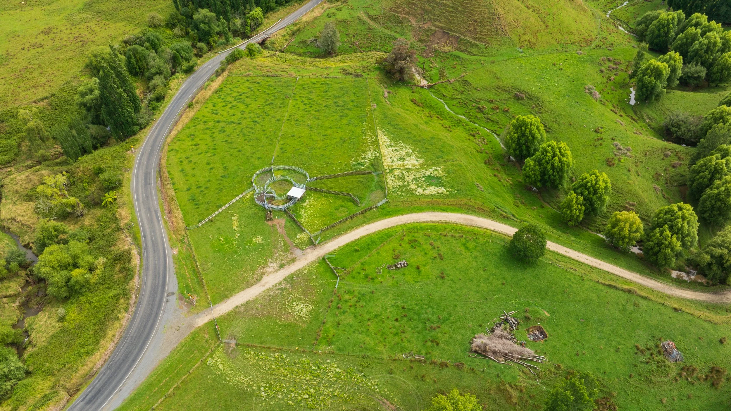 Aerial view of a green rural landscape with a winding road, a fenced circular structure, trees, and grassy fields.