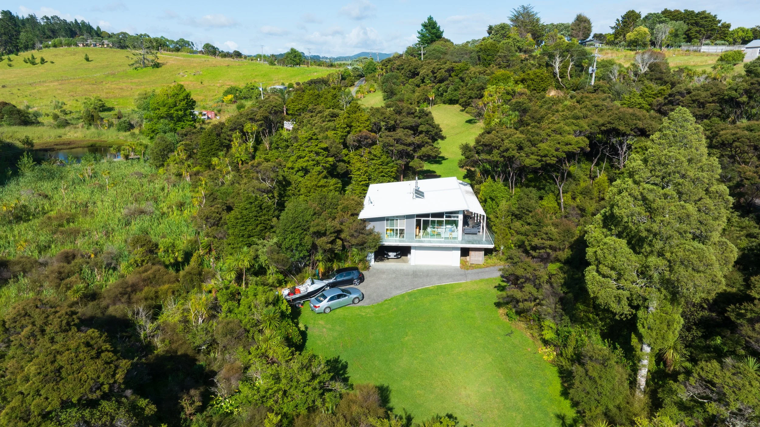 A modern white house on a hillside with a garage and driveway, surrounded by lush green trees and grass, with a scenic landscape in the background.
