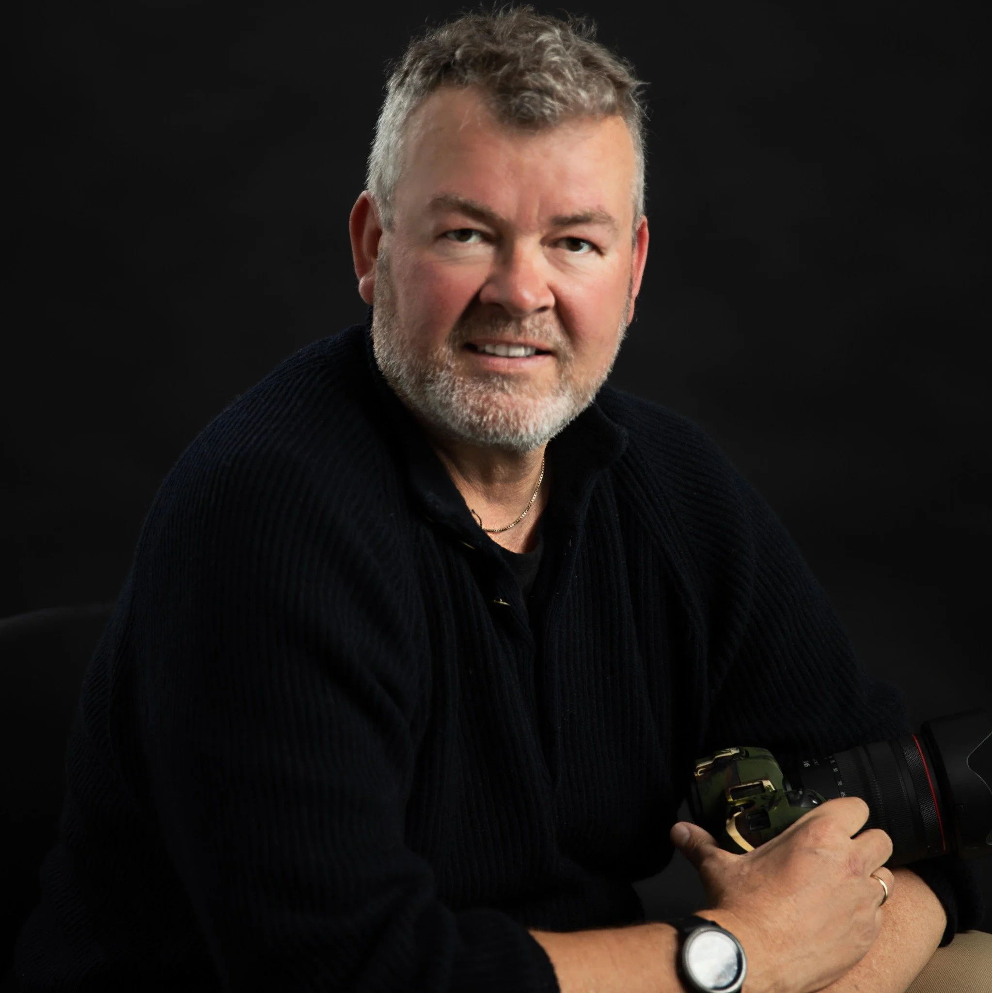 A man with gray hair and beard, wearing a black sweater, holding a camera with a large lens, against a dark background.