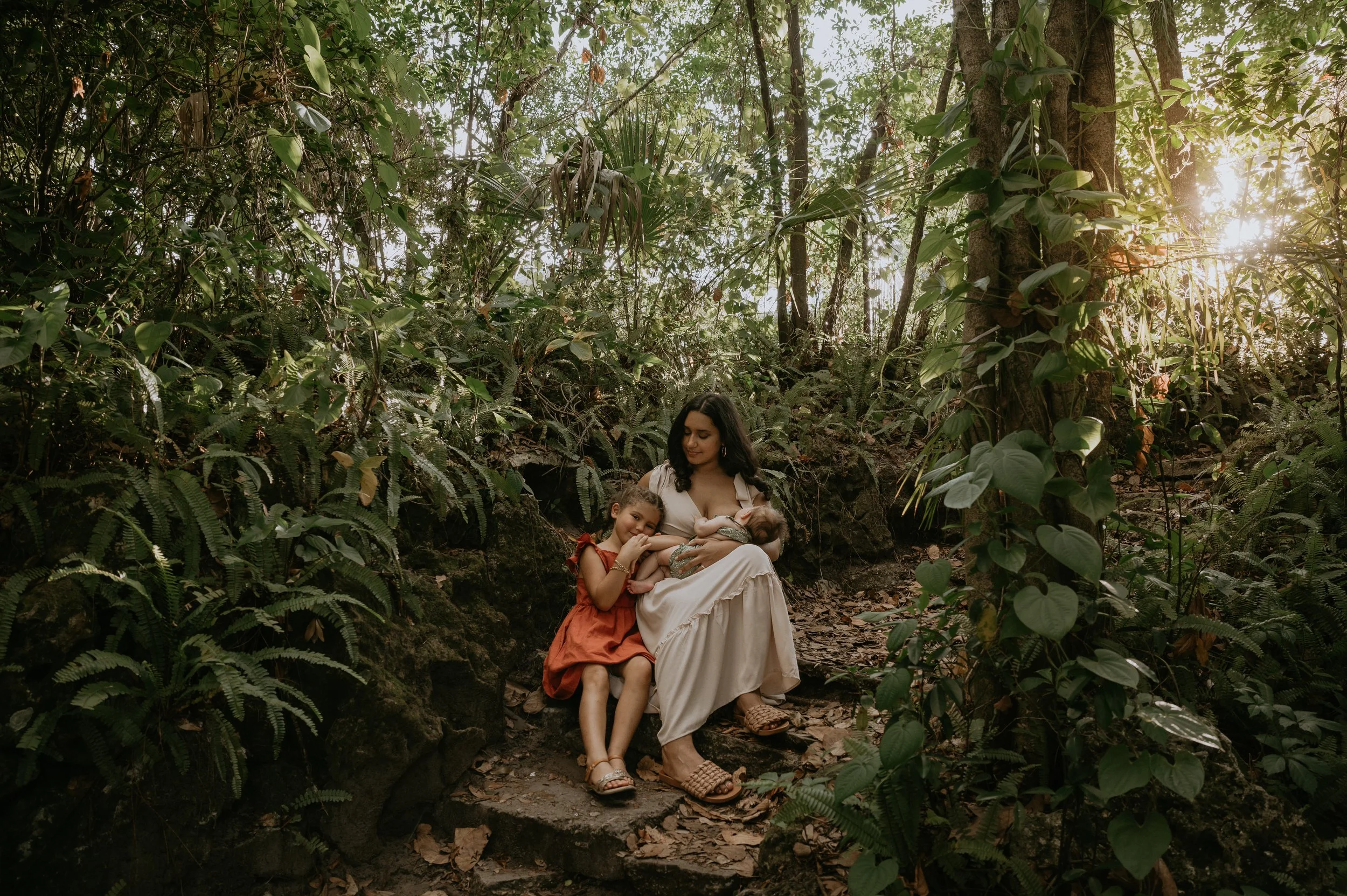 A woman is nursing an infant and hugging a young girl while sitting on a rock amongst a dense, green Florida forest, with sunlight filtering through the trees.