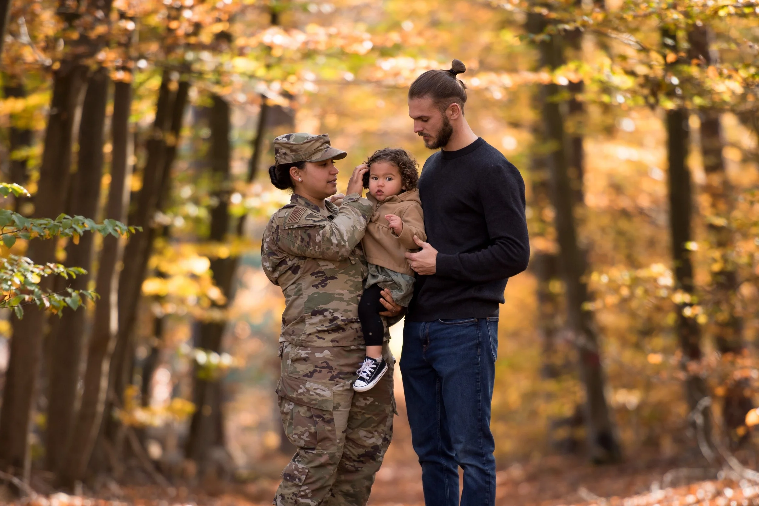 A woman in military uniform holding a young child who is touching her face, while a man stands close beside them in a wooded area with autumn leaves.