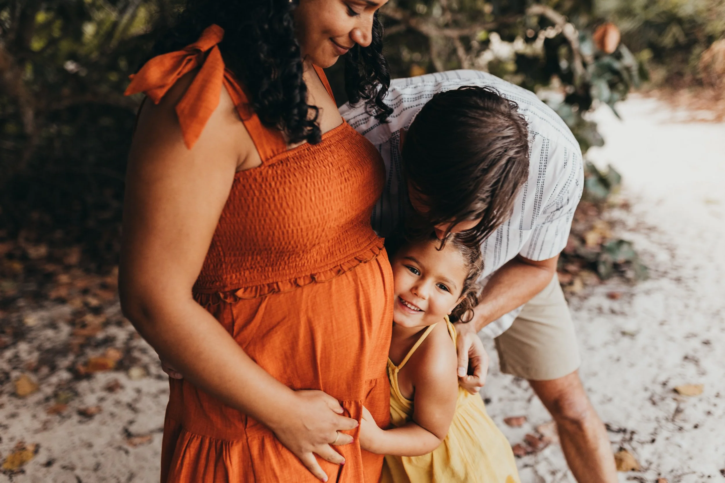 A pregnant woman, her husband  and their young daughter are embracing outdoors, smiling, near a river with trees in the background.