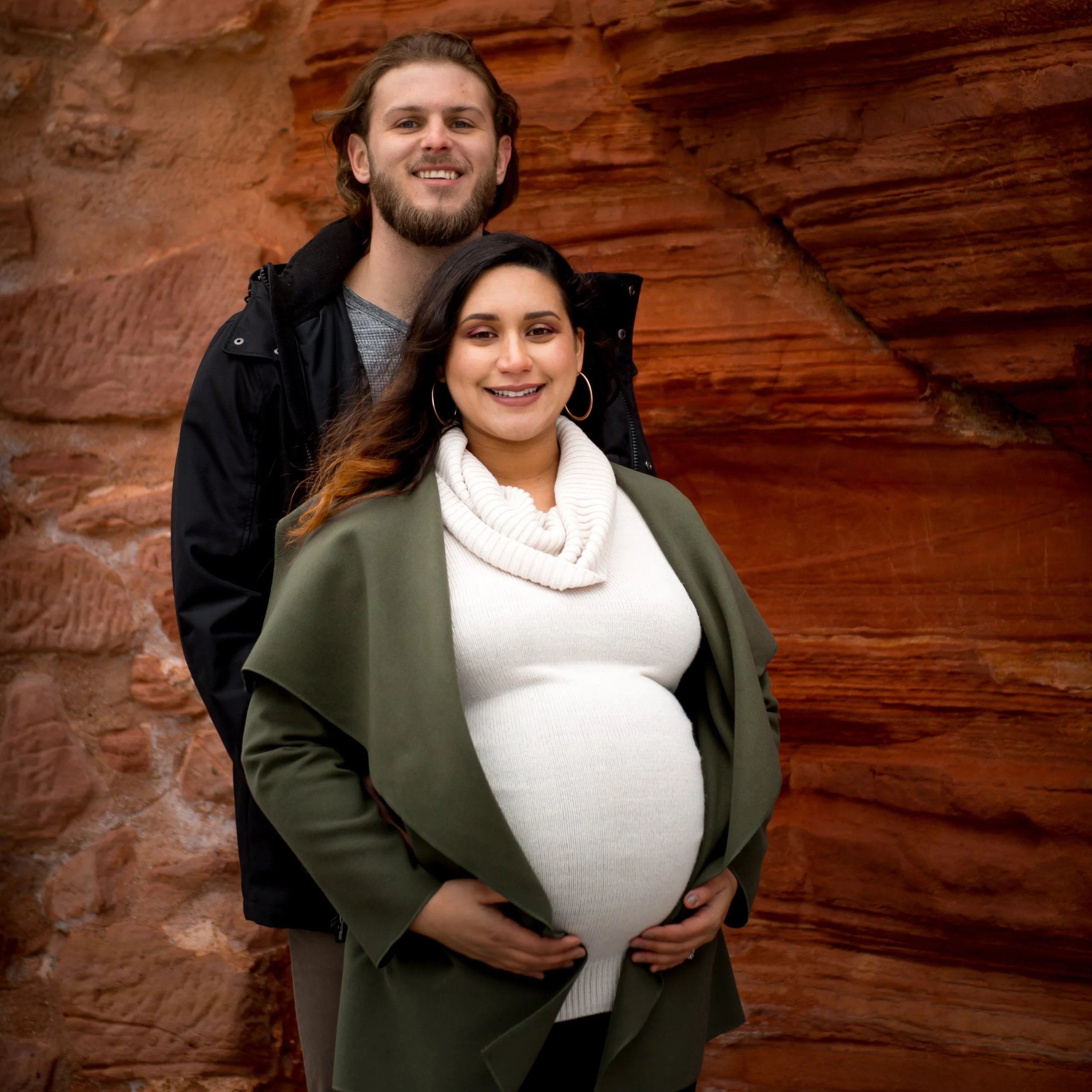 A young man and pregnant woman smiling outdoors against a red castle wall in Germany. The man has long, wavy hair and a beard, wearing a black jacket over a gray shirt. The woman has dark, wavy hair, wearing a white turtleneck and a green jacket.