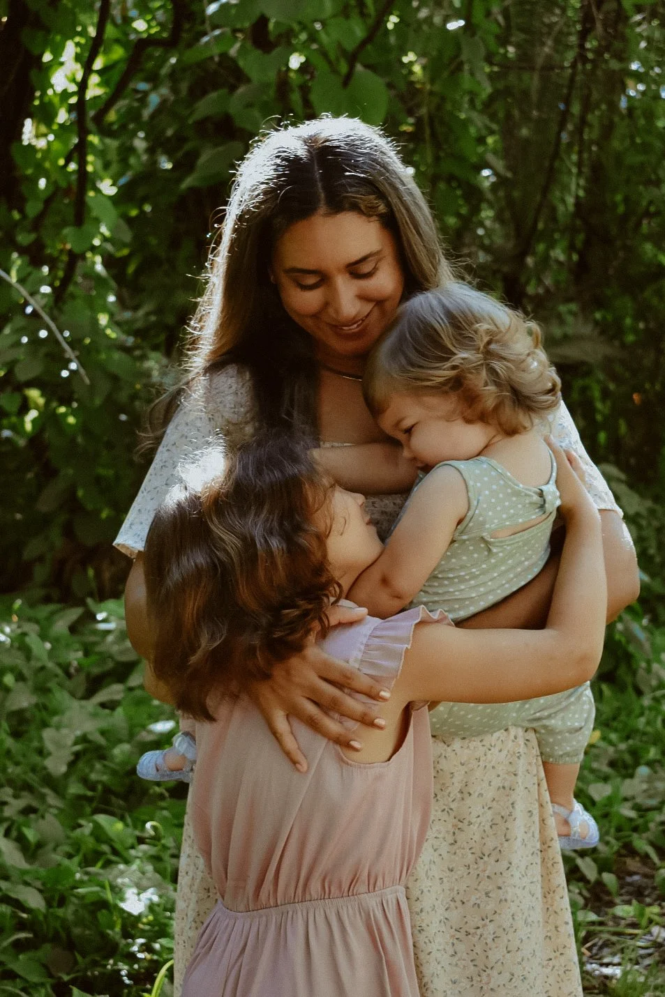 A woman with long brown hair hugging and smiling at two young girls with curly hair, one girl with brown hair wearing a pink dress and the other girl with blonde hair wearing a green polka-dot outfit, in a green, leafy outdoor setting.