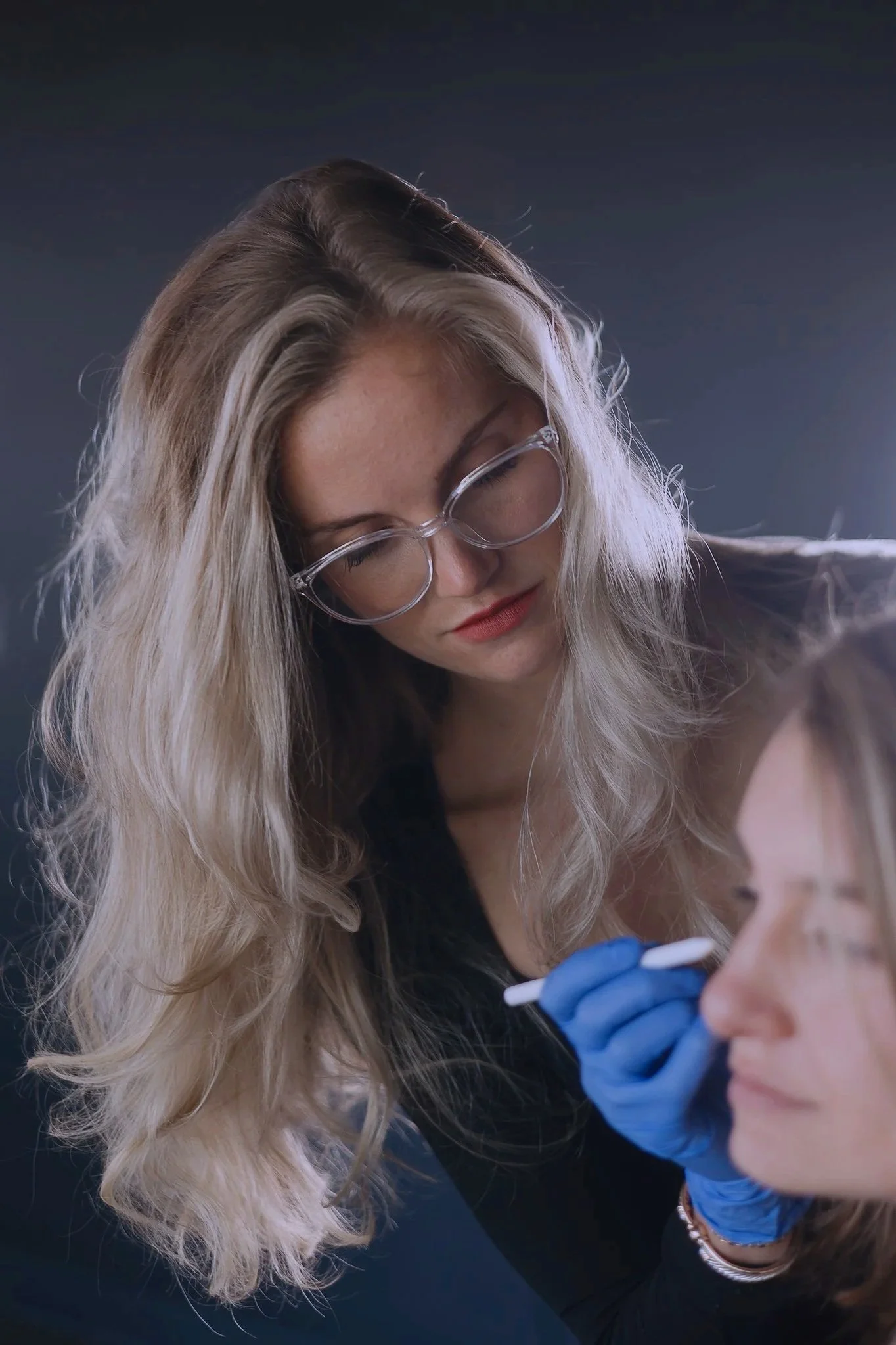 A woman with long blonde hair, wearing glasses, performs a beauty or cosmetic procedure on another woman with glasses, using a small tool and wearing blue gloves.